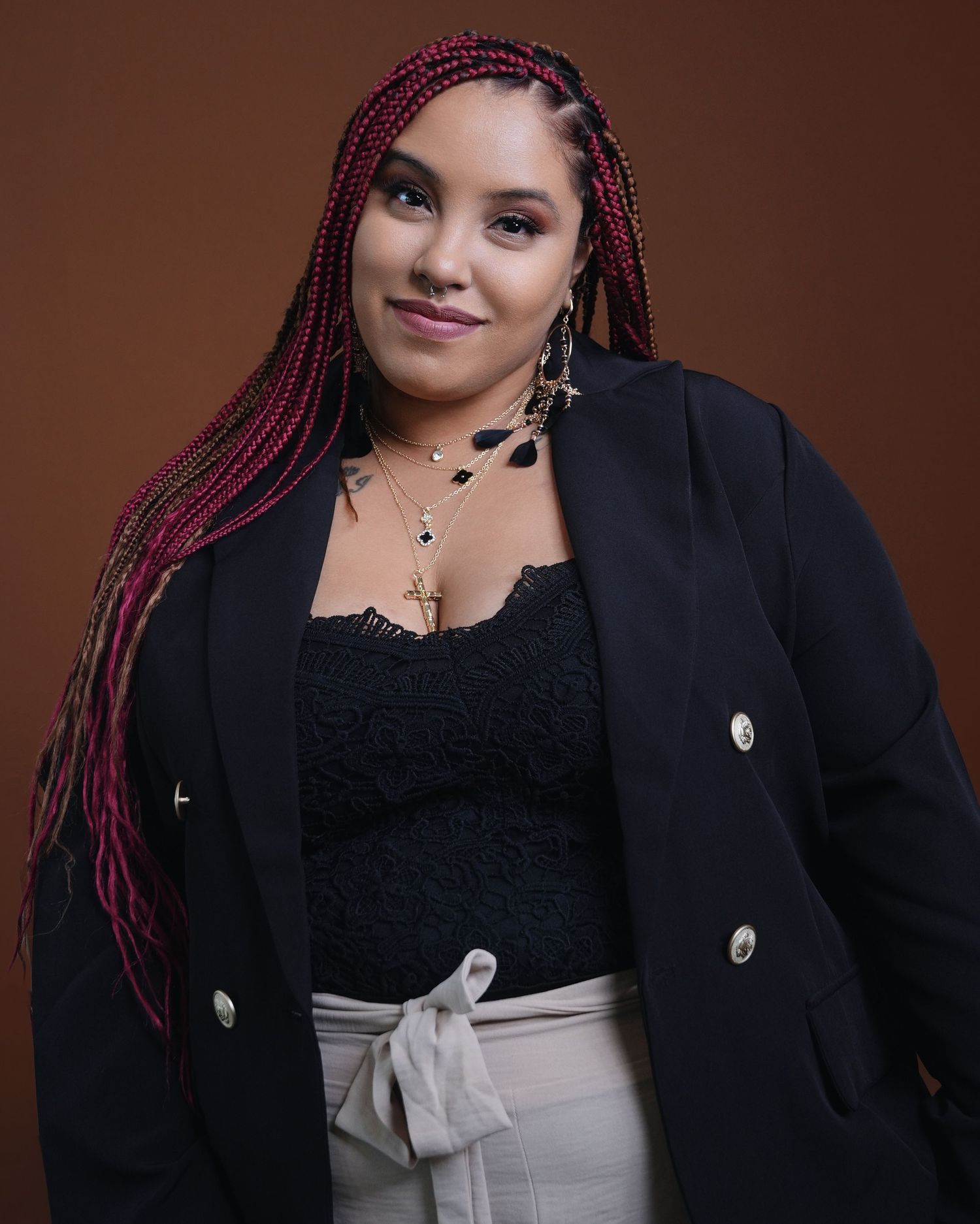 Professional portrait of performer with burgundy braids wearing black blazer against brown backdrop.
