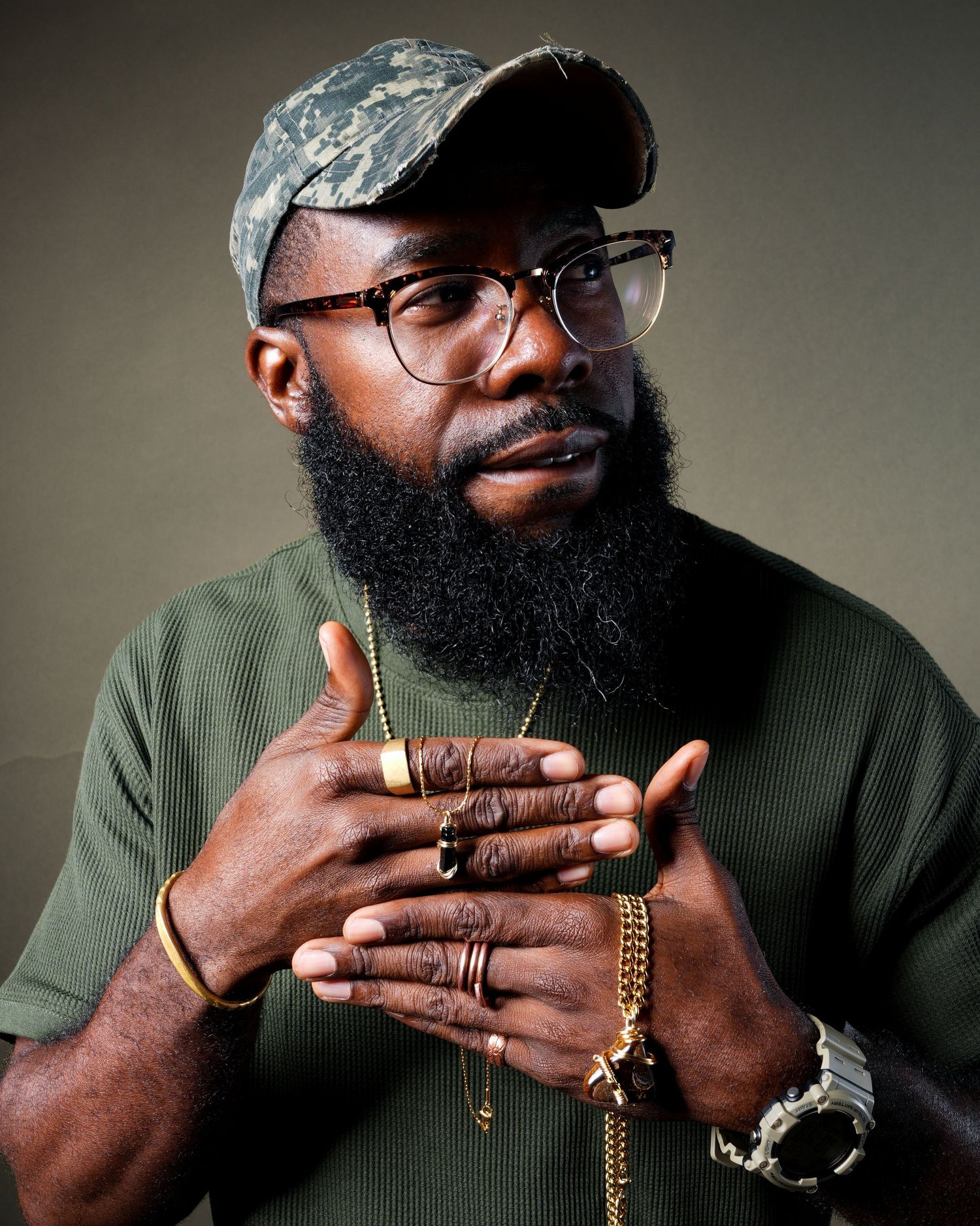 Portrait of someone wearing glasses and a blue cap with a full beard, wearing green, hands clasped with jewelry.