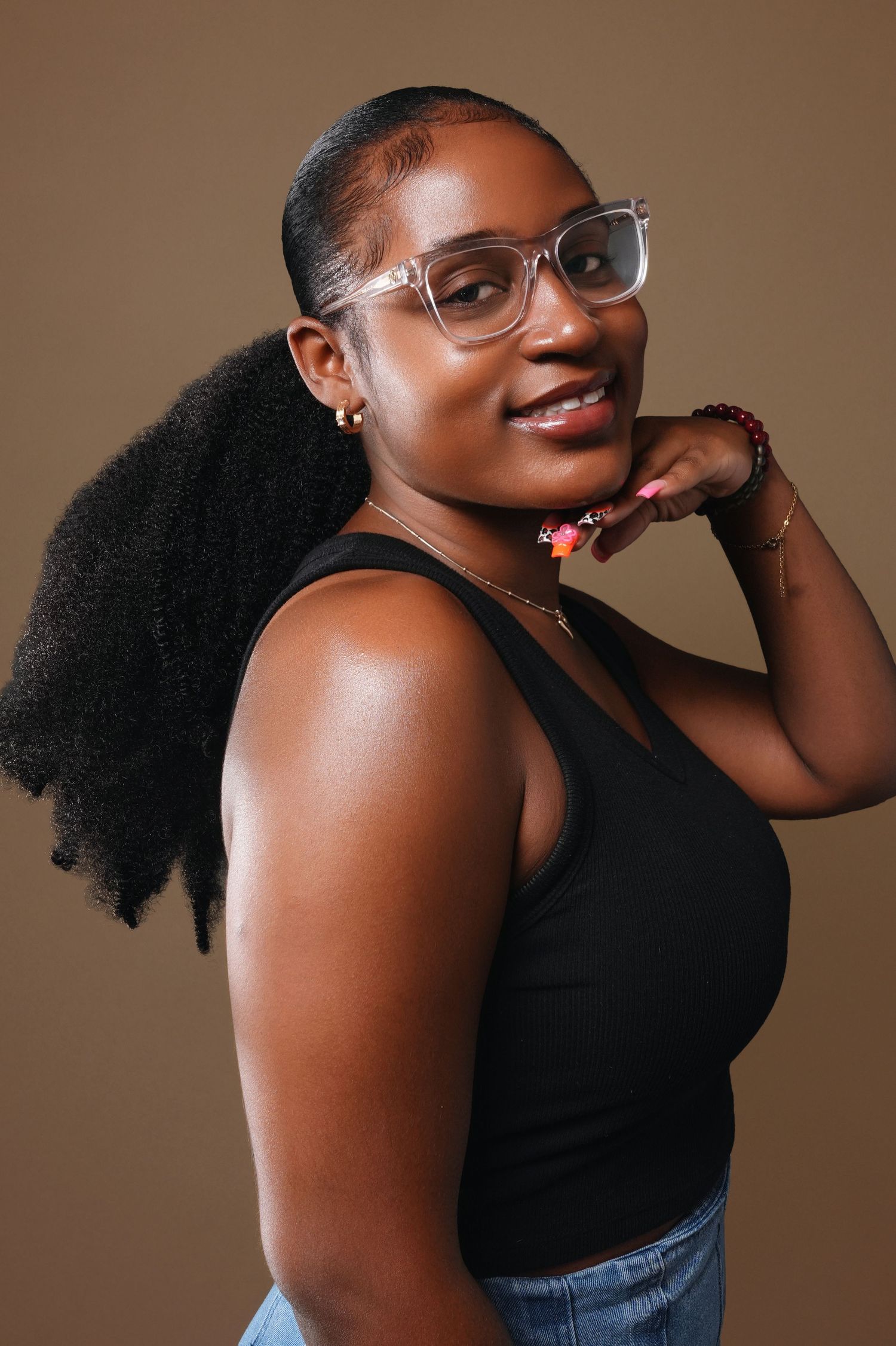 Studio portrait of a person with natural hair wearing glasses and black tank top against brown background.