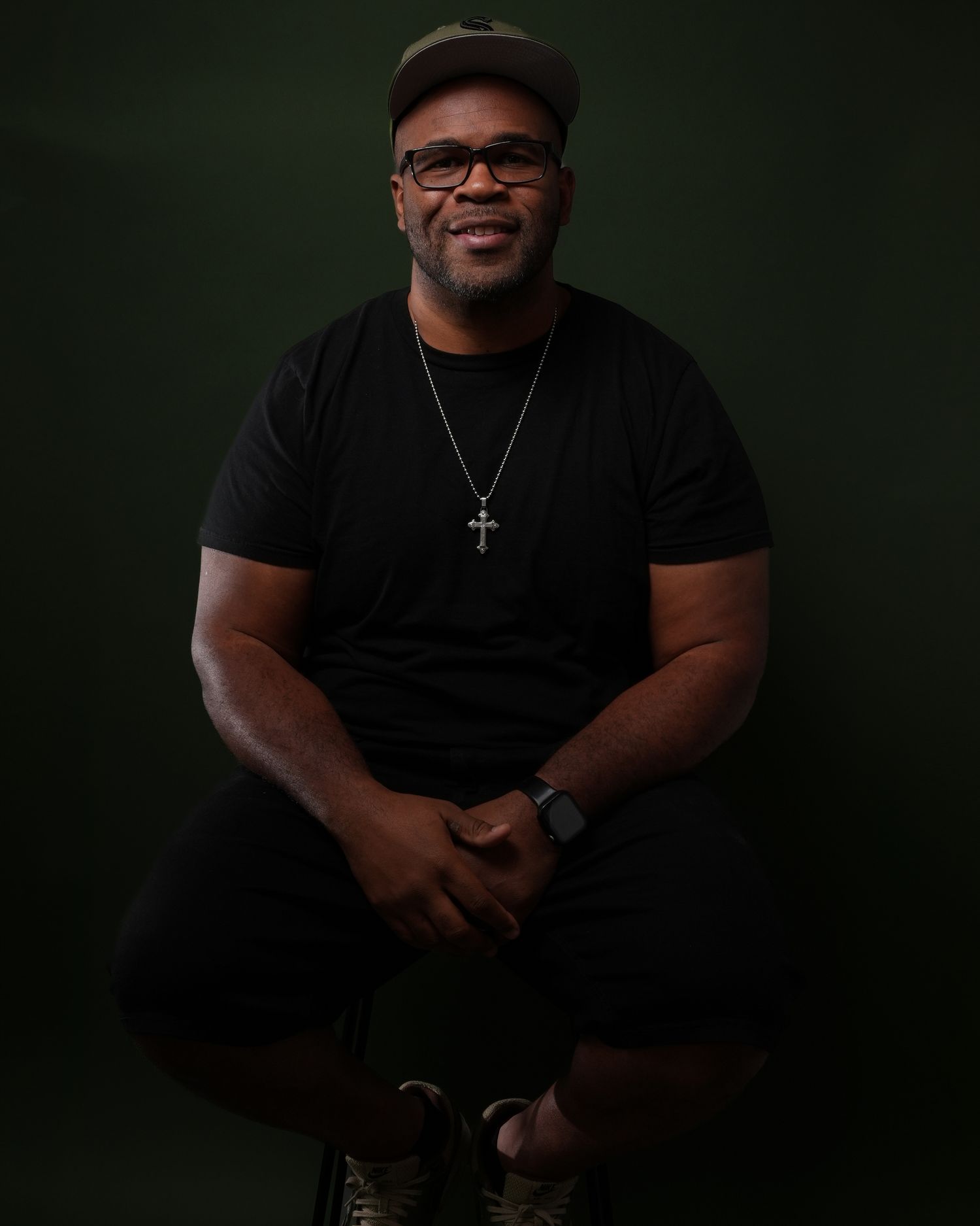Person wearing glasses, baseball cap and cross necklace smiles during professional studio photoshoot.