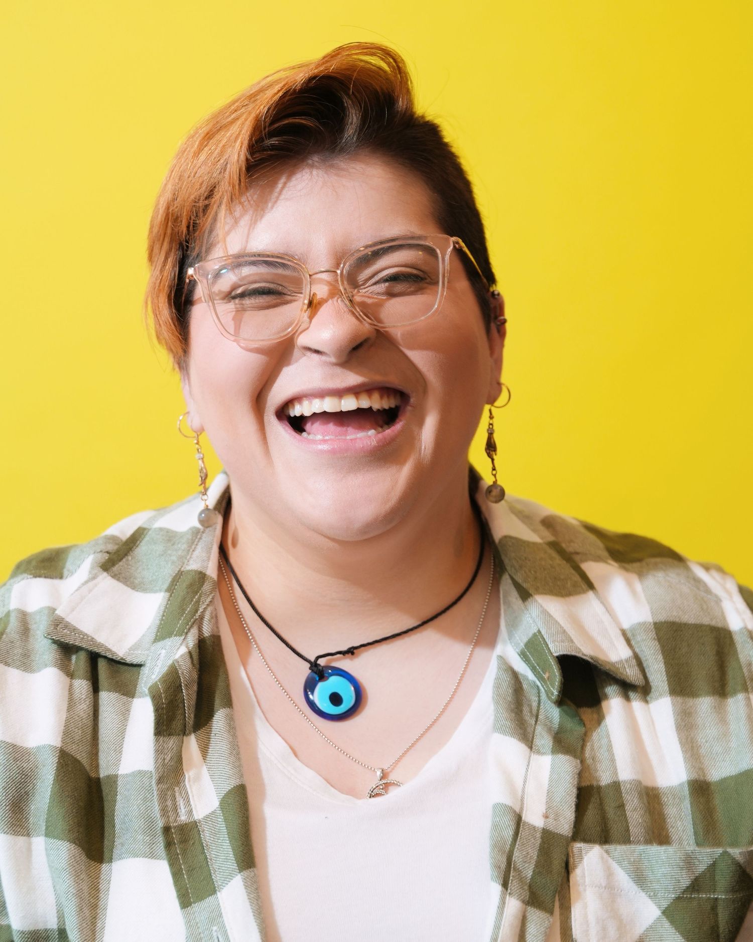 Joyful portrait in plaid shirt and glasses with blue pendant necklace against yellow background.