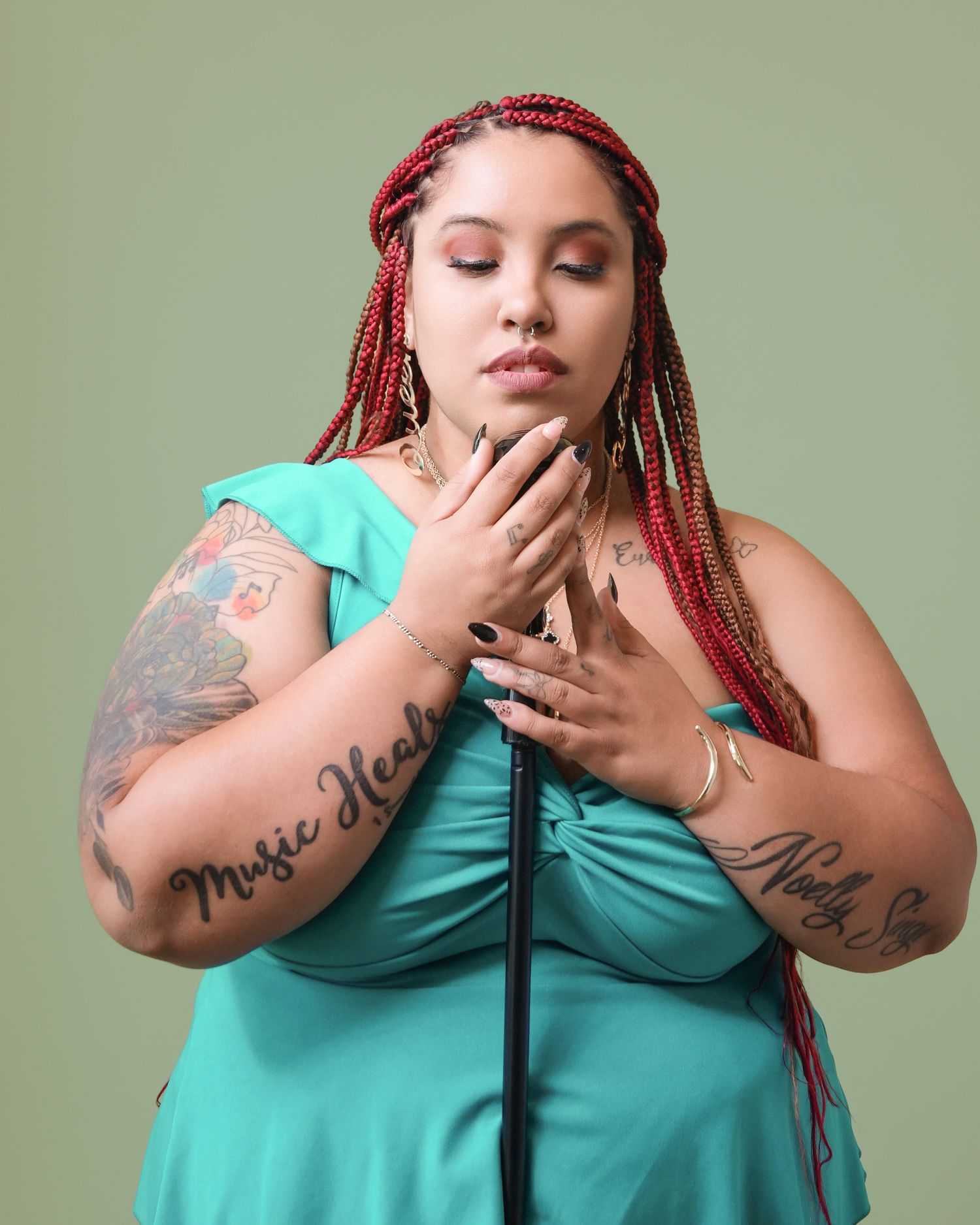 Artistic portrait of performer with red braids in turquoise dress showing tattoos against green backdrop.
