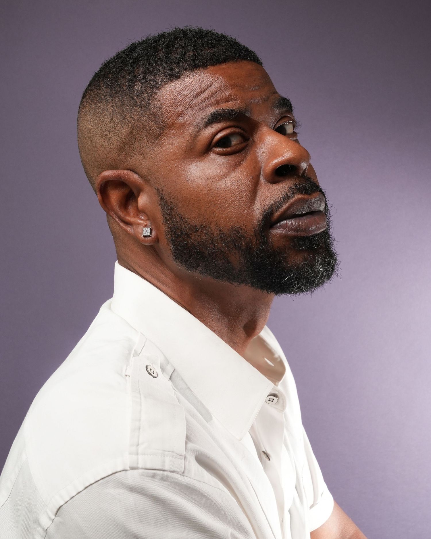 Side profile portrait of man wearing white button-up shirt with an earring against purple background.