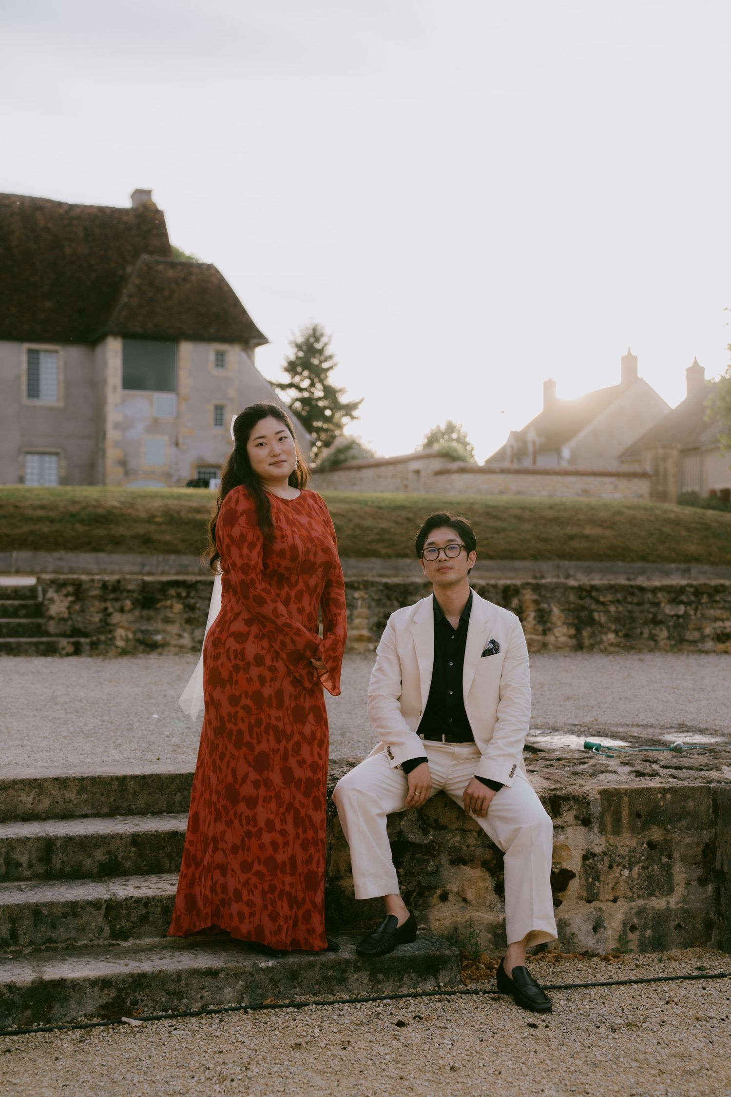 Couple poses on stone steps outdoors with one person wearing a striking red dress and another in white attire.