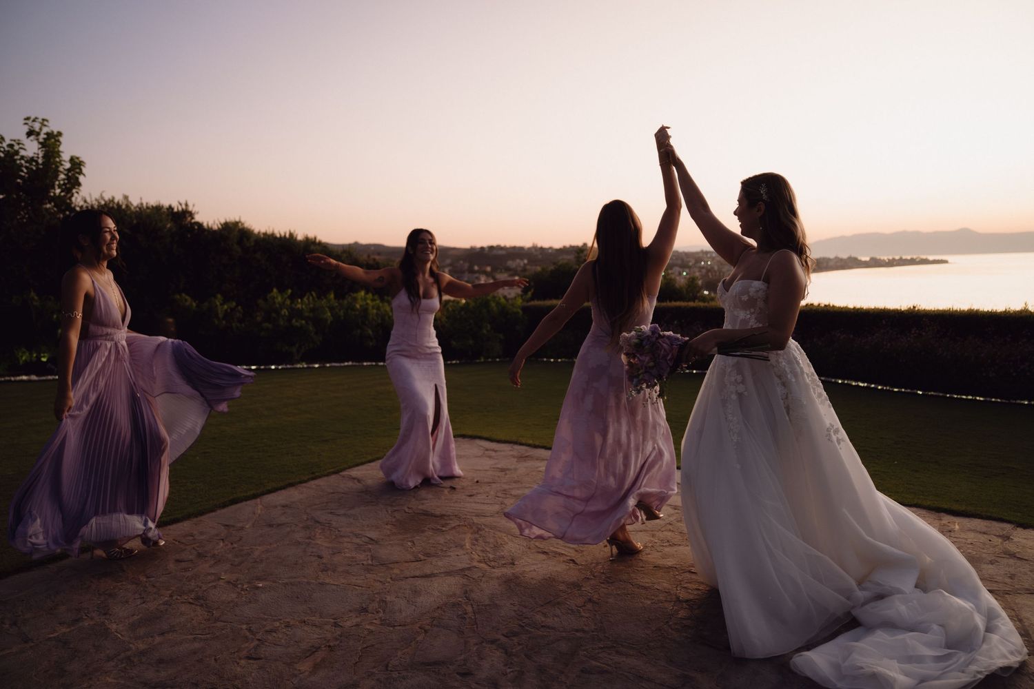 Wedding party in lavender dresses poses at sunset on a scenic hilltop overlooking water.