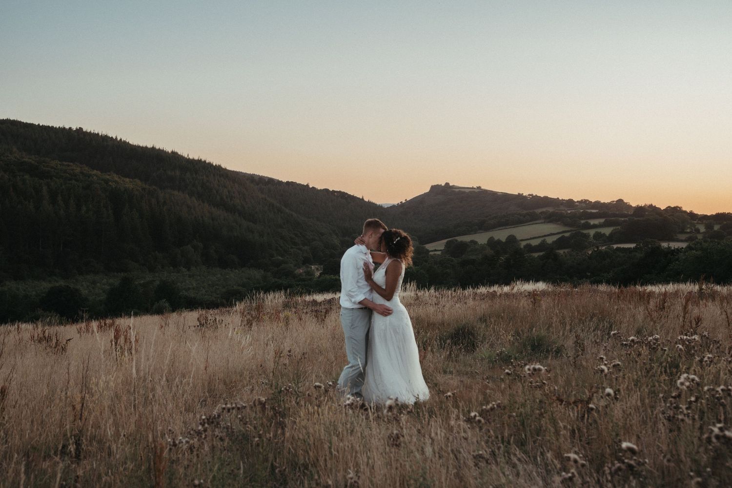 Wedding couple embraces in a serene mountain field at golden hour.