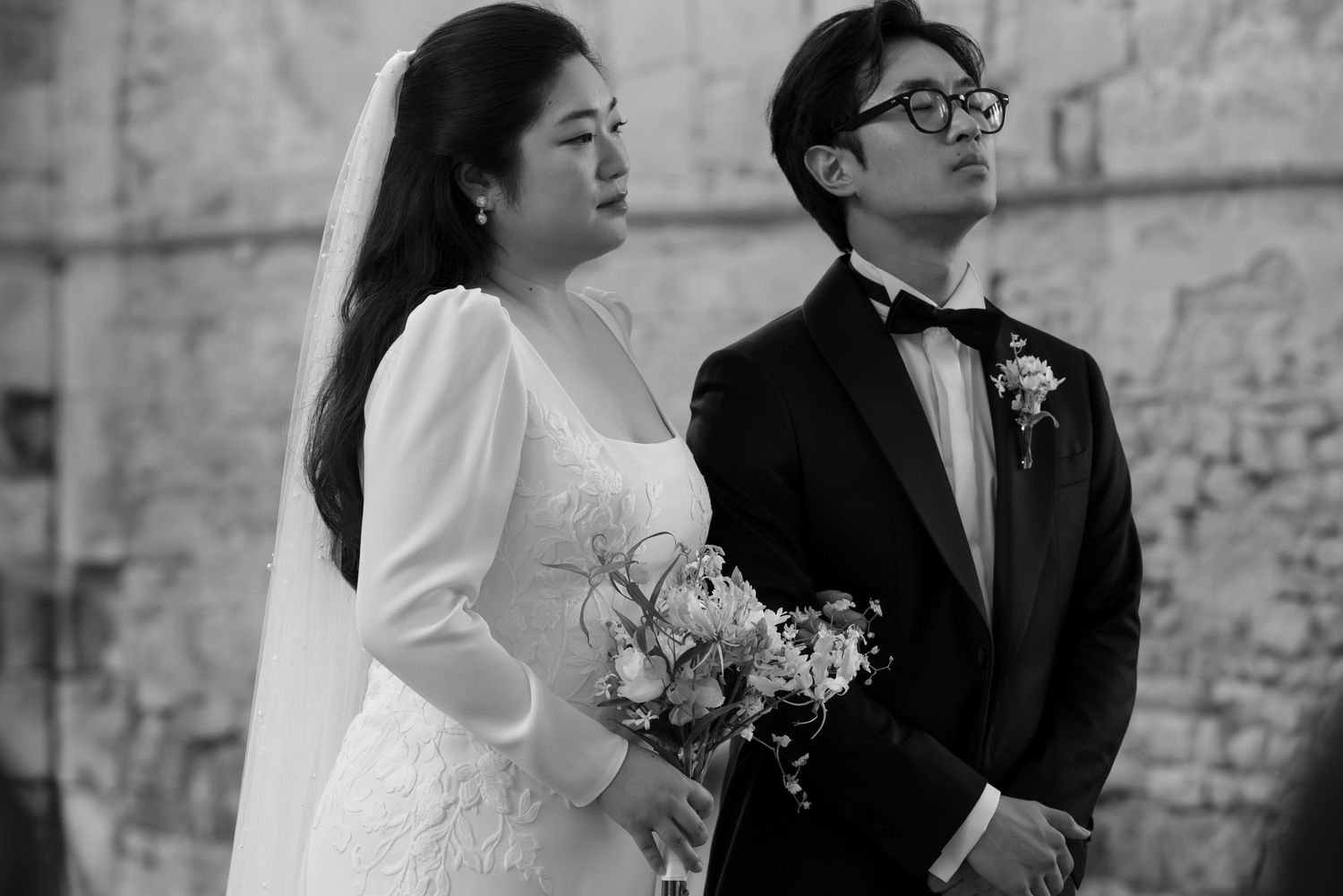 A couple in formal wedding attire poses together against a stone wall backdrop during their wedding ceremony.