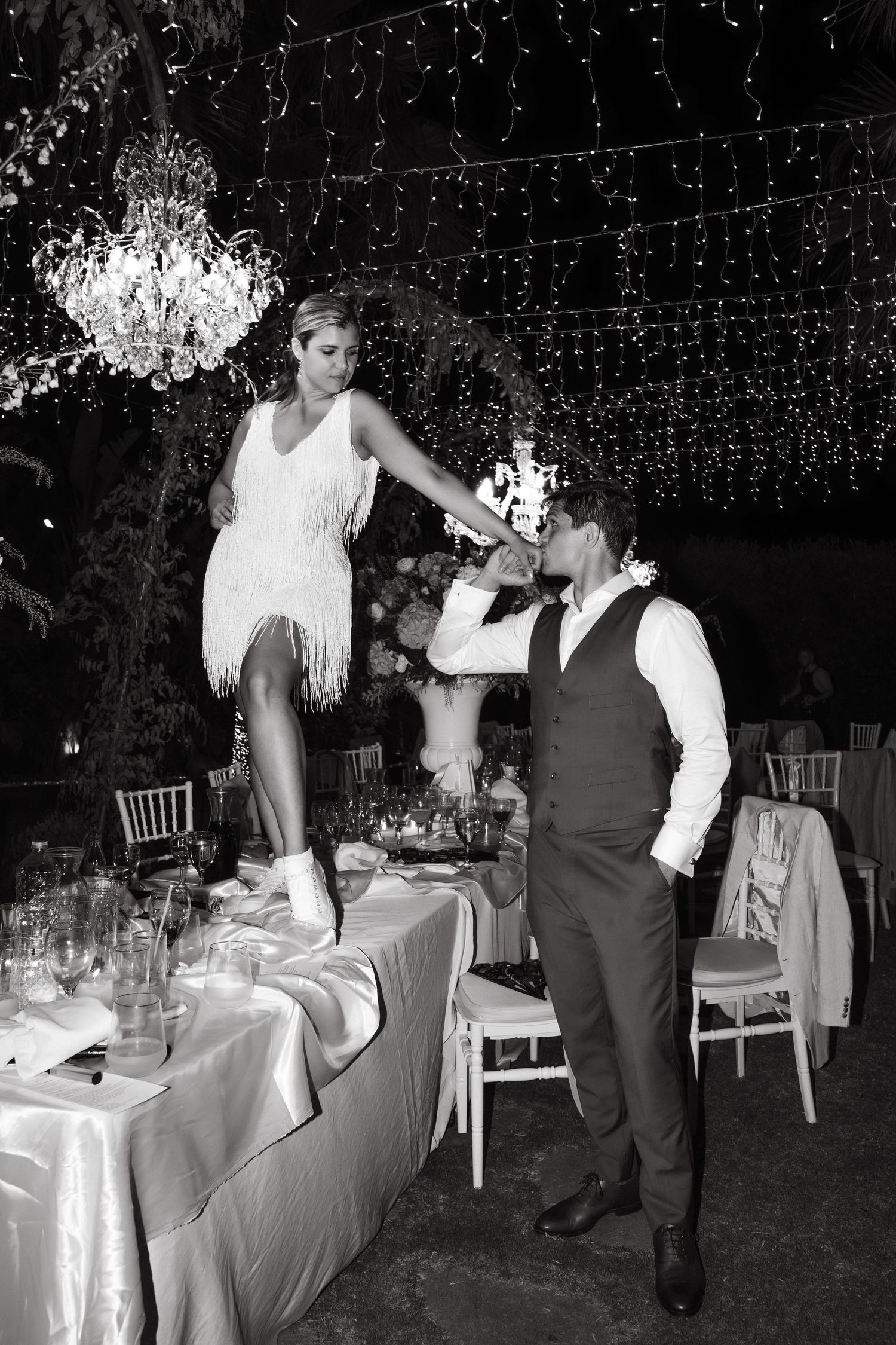 Black and white photo of wedding guests mingling under string lights at an outdoor reception.