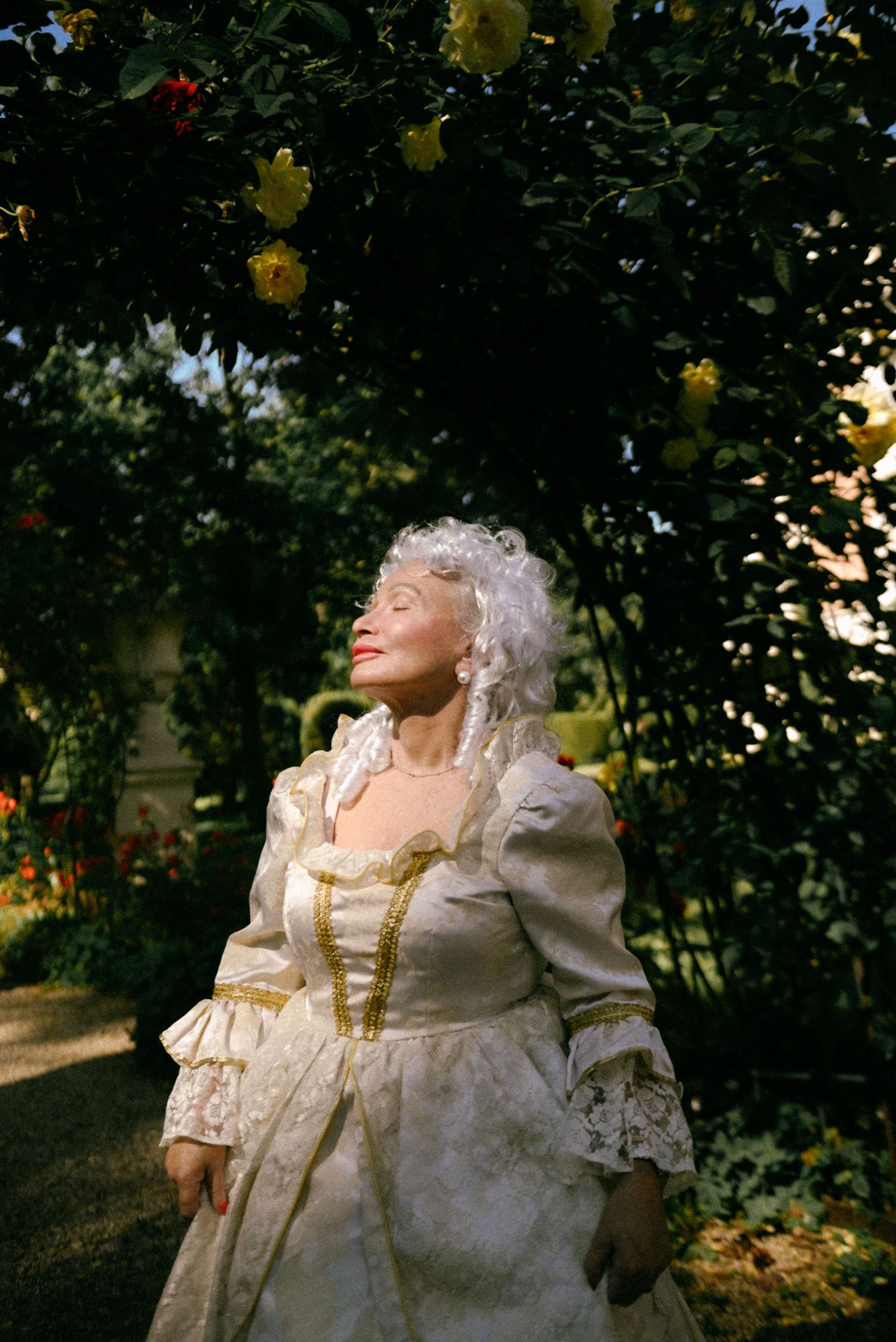 Person wearing an ornate white and gold baroque or rococo style period costume poses outdoors among trees and foliage.