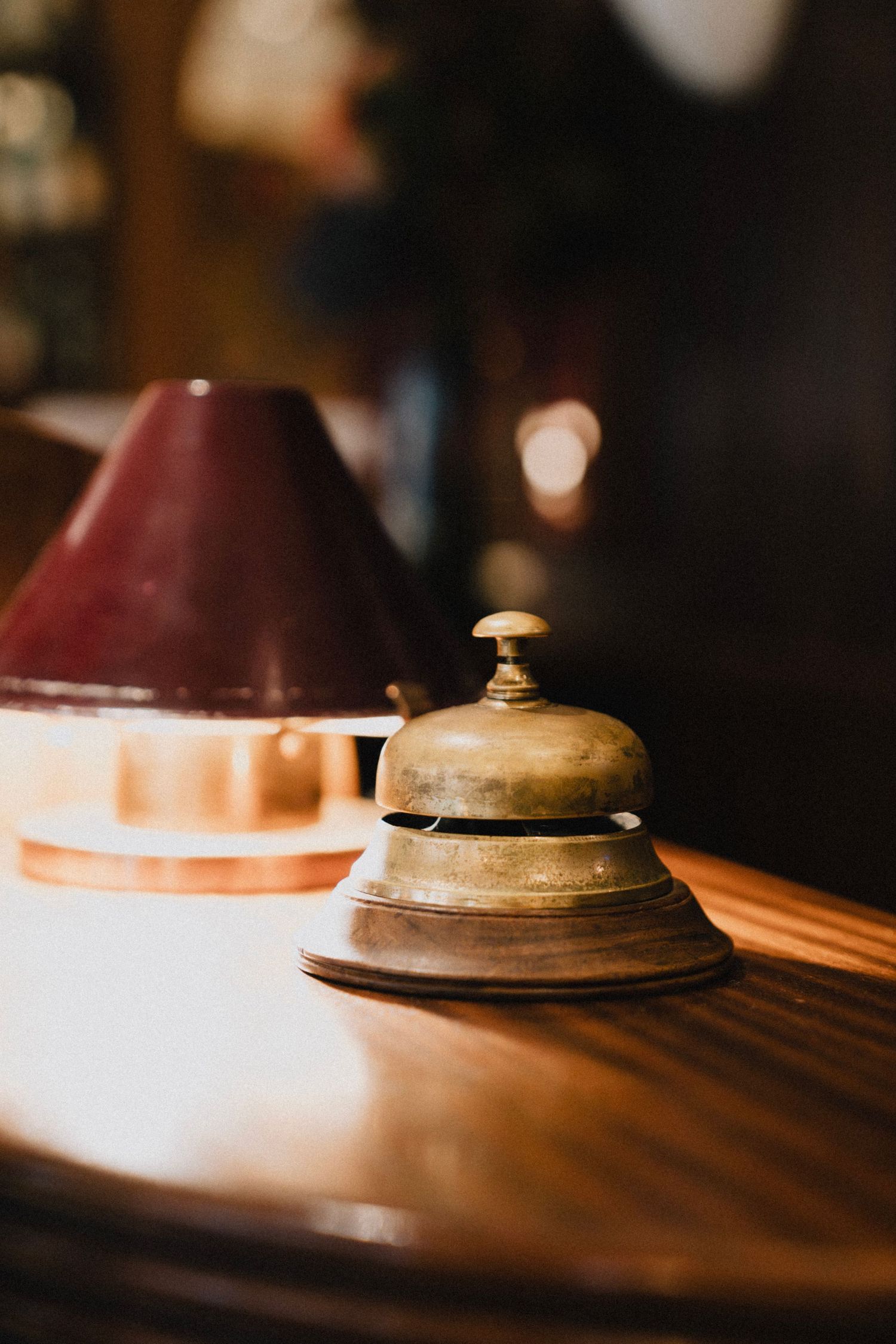 Vintage brass service bell sits on polished wooden surface in warm lighting with bokeh background.