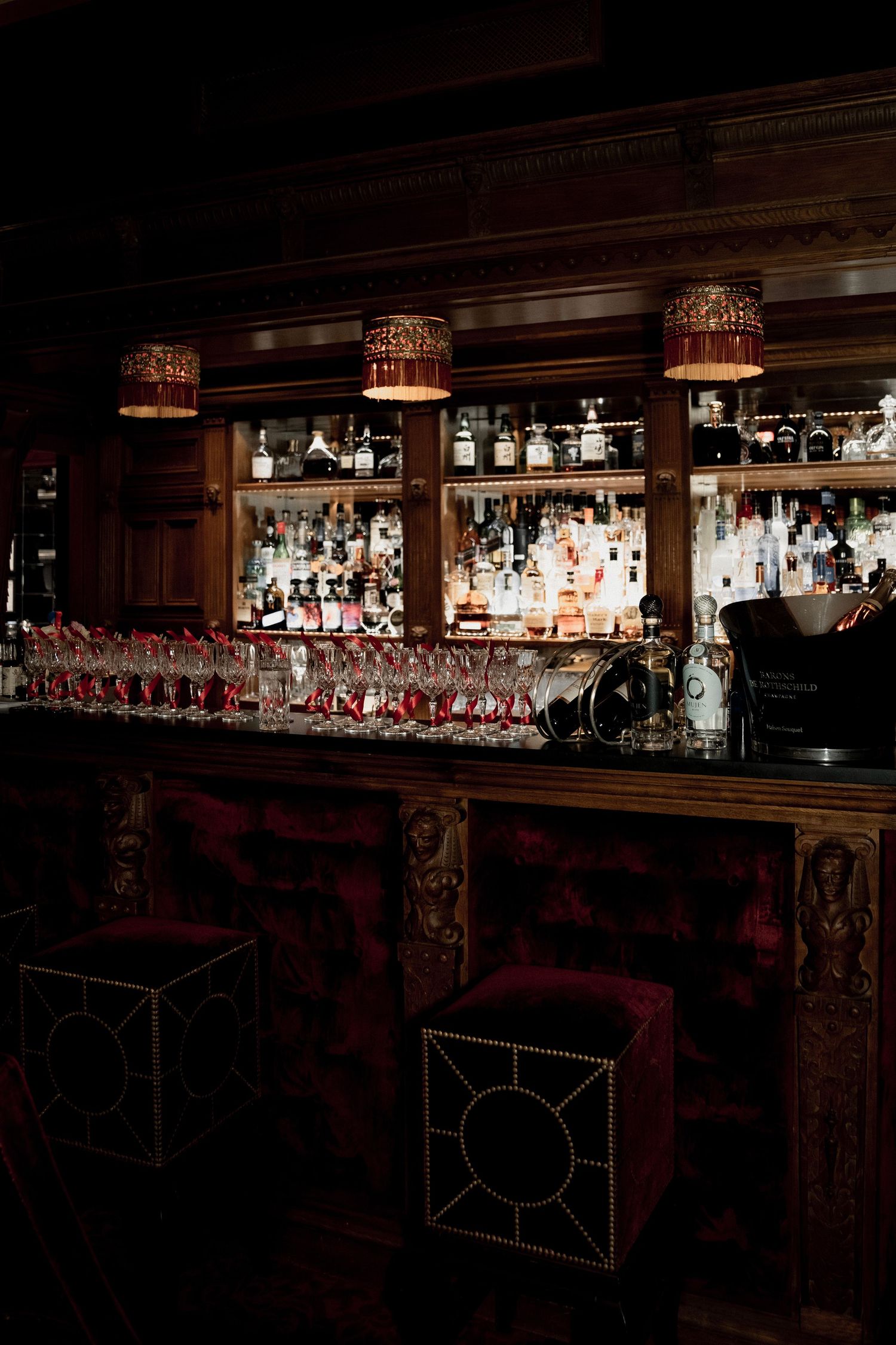 Dark wooden bar interior with illuminated liquor bottles on shelves and hanging wine glasses in elegant ambient lighting.
