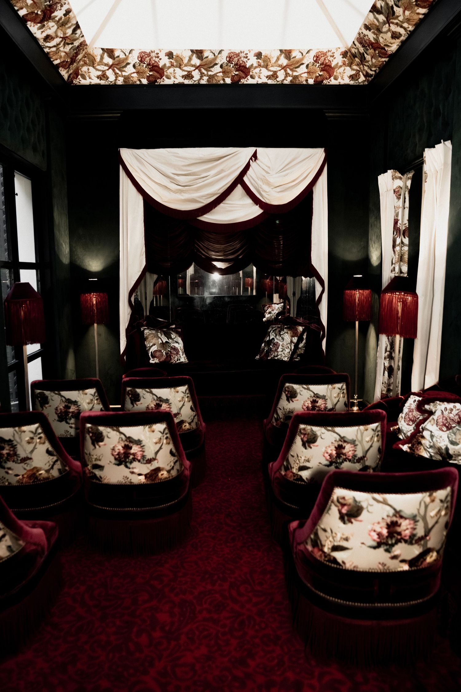 Luxurious dining room with floral upholstered booths, white drapes, and red carpeting in dim lighting.