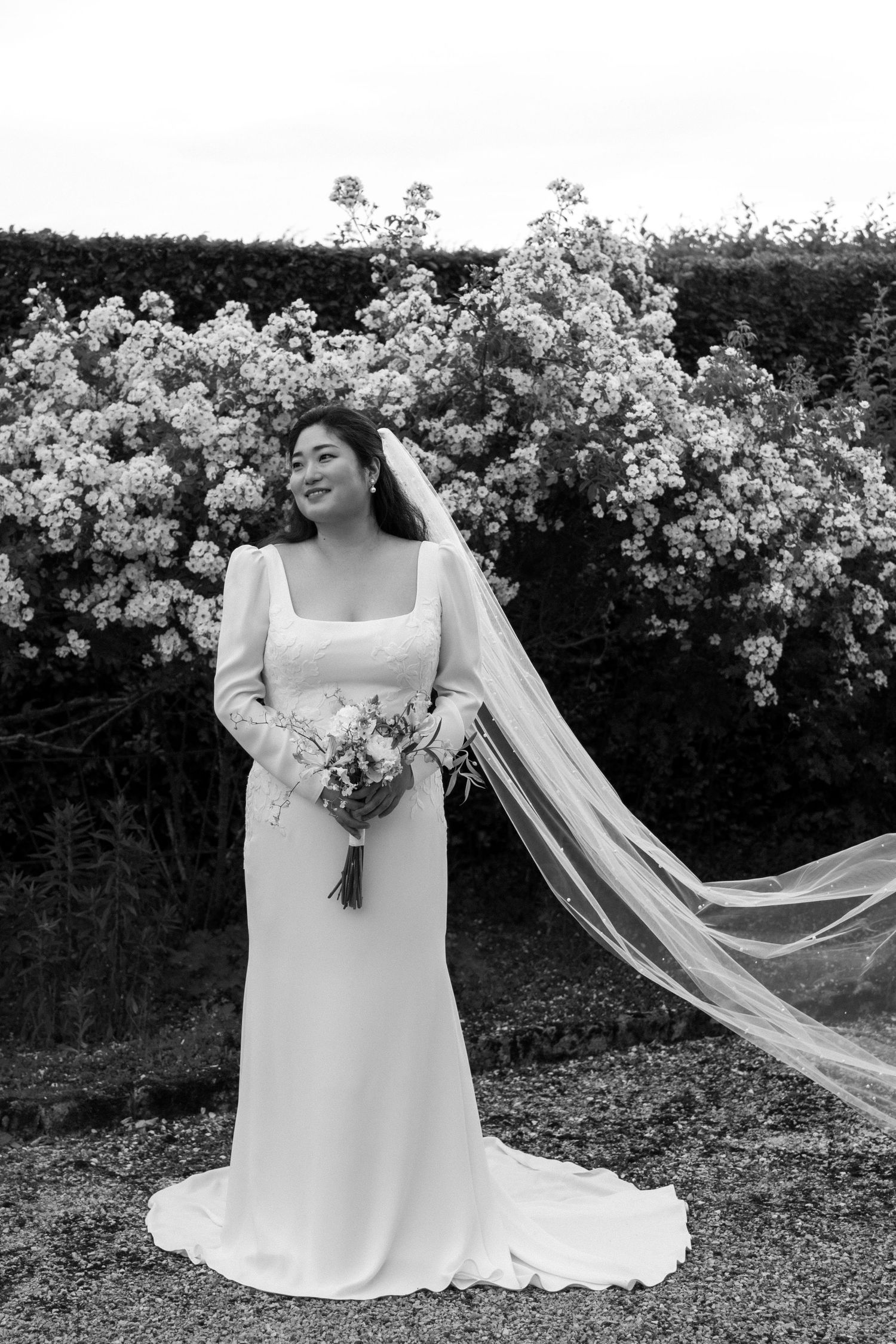 A bride in a flowing white wedding dress poses with a bouquet against a backdrop of blooming white flowers.