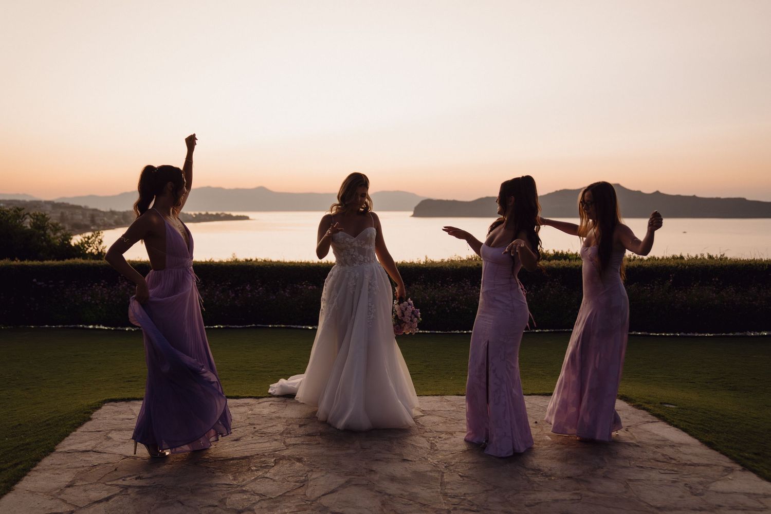 Wedding party silhouettes sit on a wall at sunset overlooking a scenic mountain and lake view.