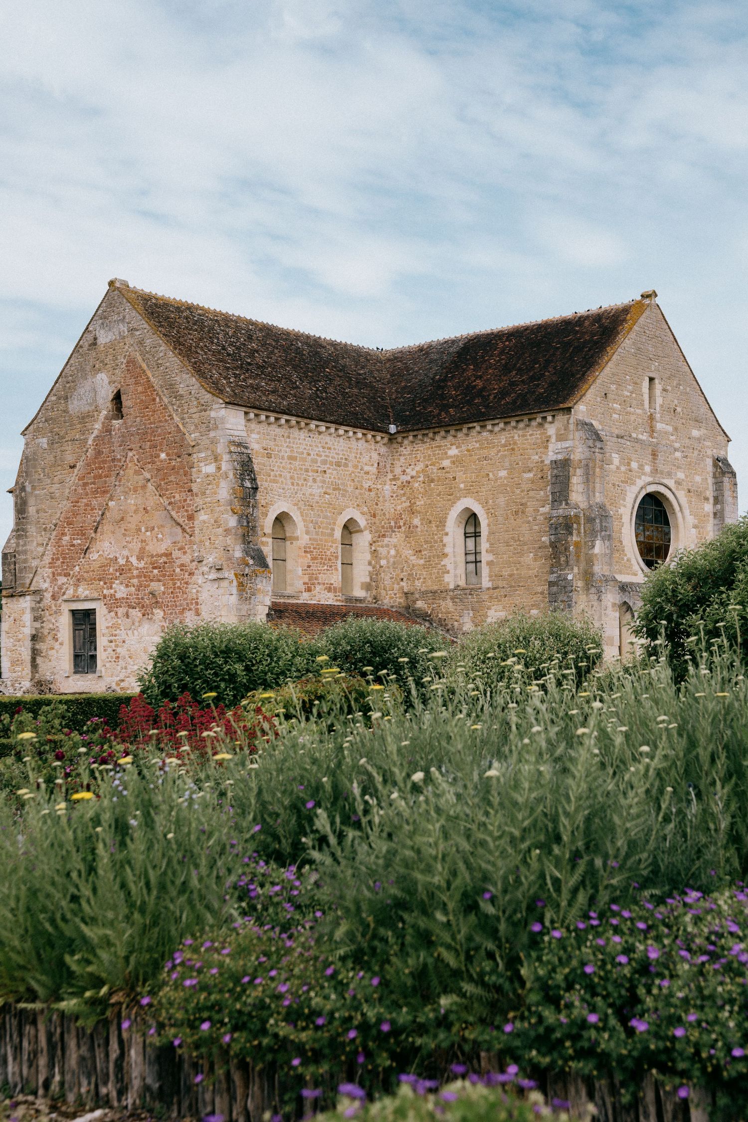 Historic stone abbey buildings with arched windows stand behind a wild garden of tall flowering plants under a blue sky.