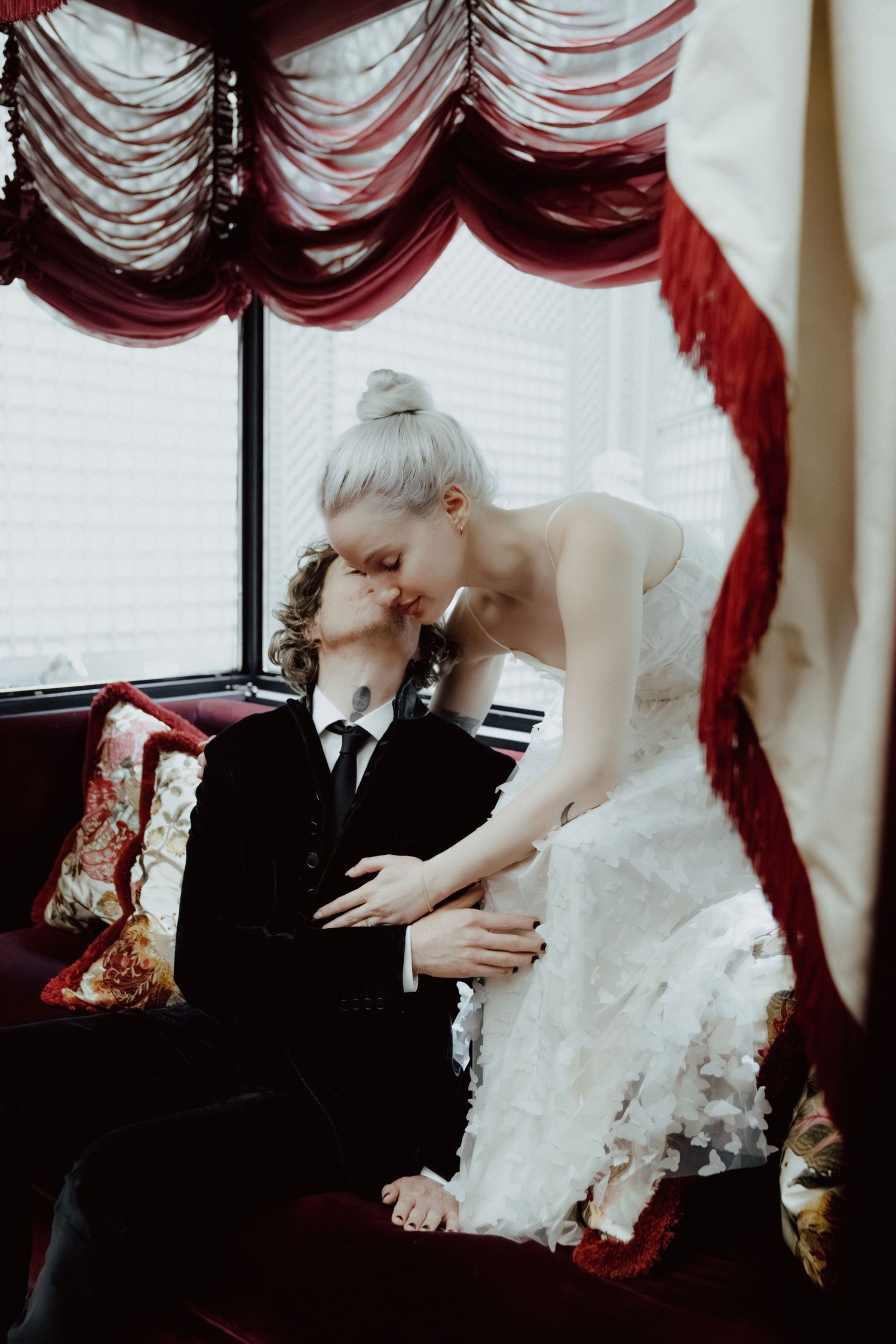 A romantic wedding scene with a couple sharing tender moments by a window with red curtains.