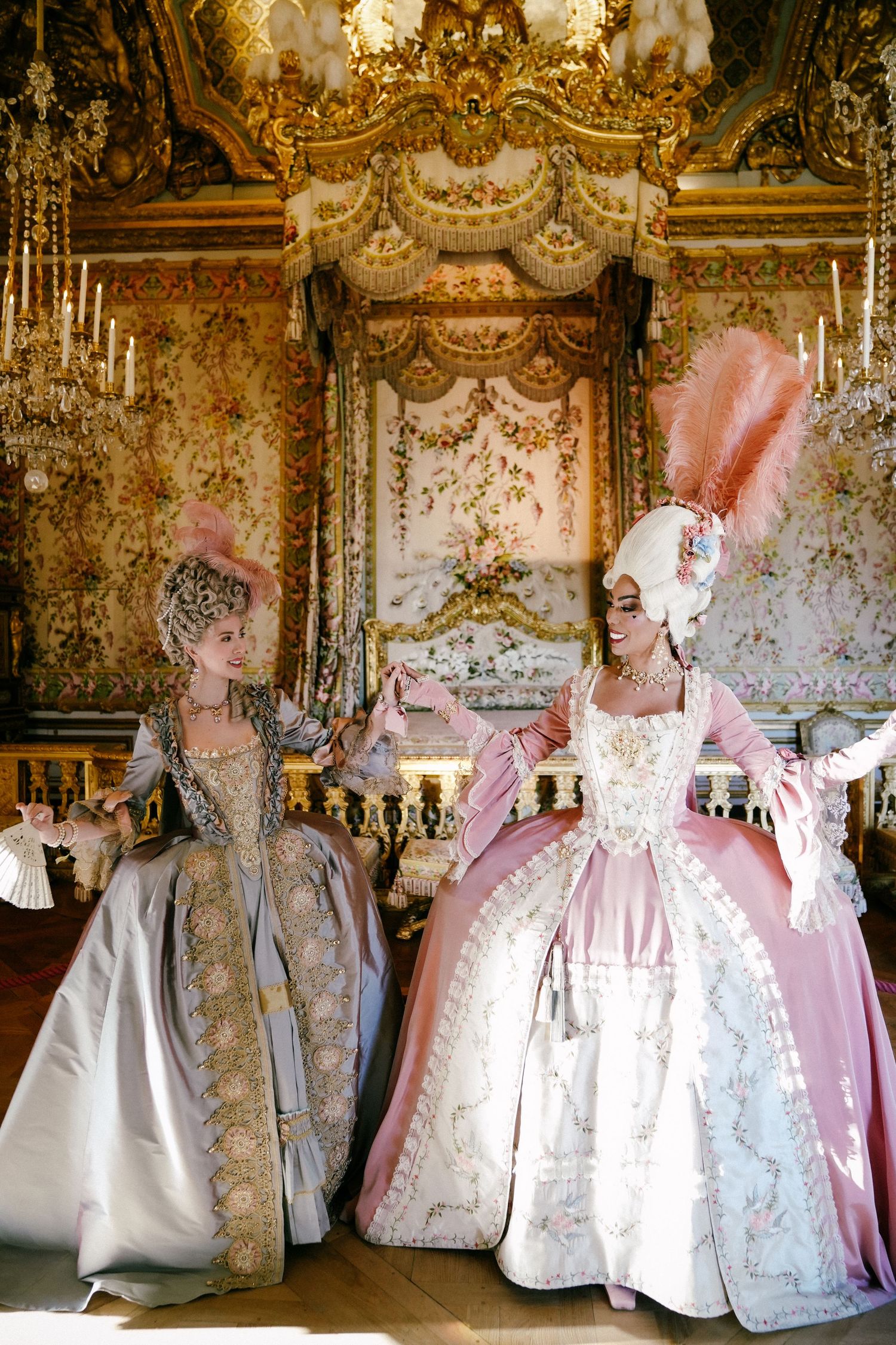 Two women pose in Marie Antionette's bedroom with period costumes featuring white and pink dresses displayed against gilded walls.