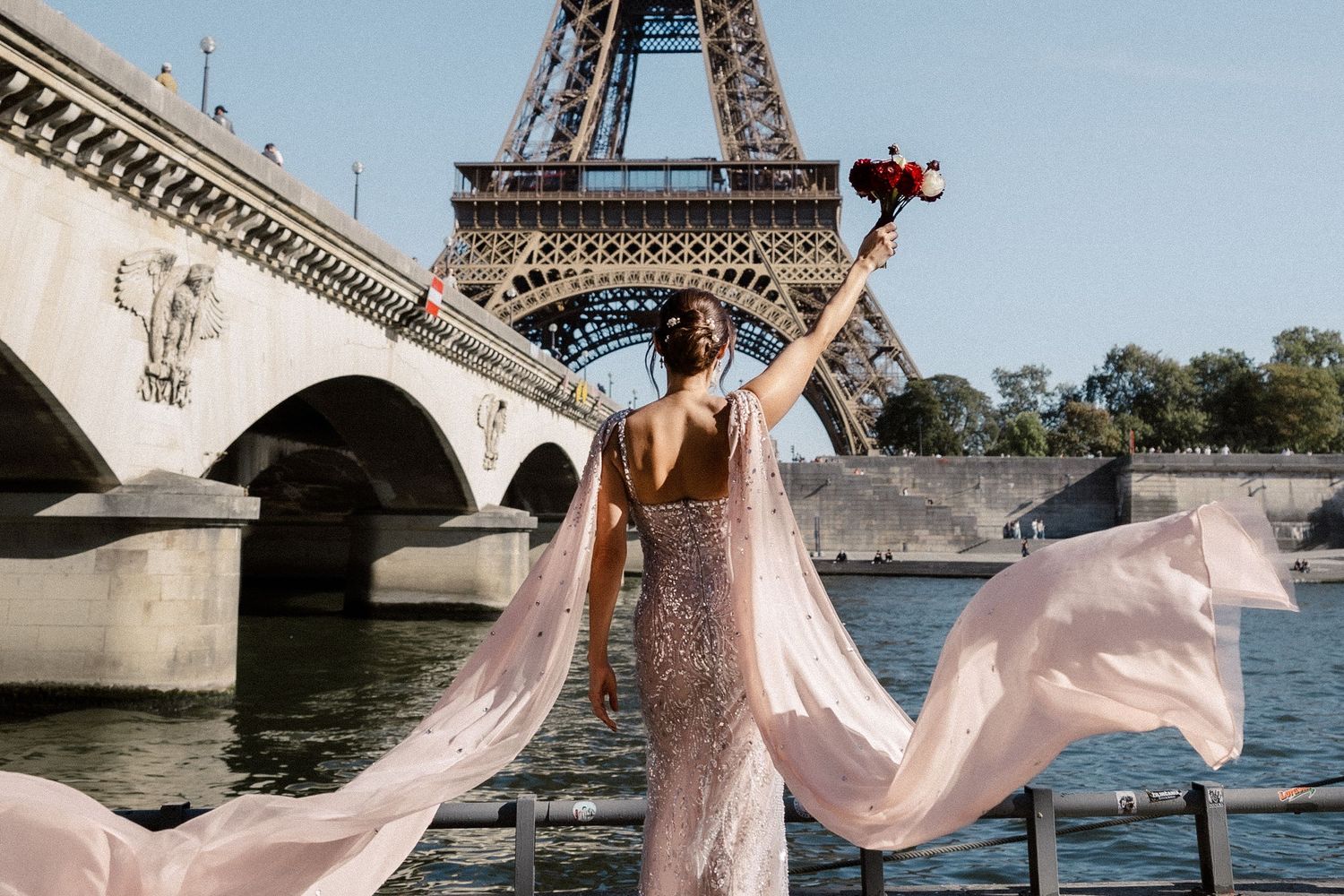 Woman in pink gown poses with bouquet by Eiffel Tower and Seine River on sunny day.