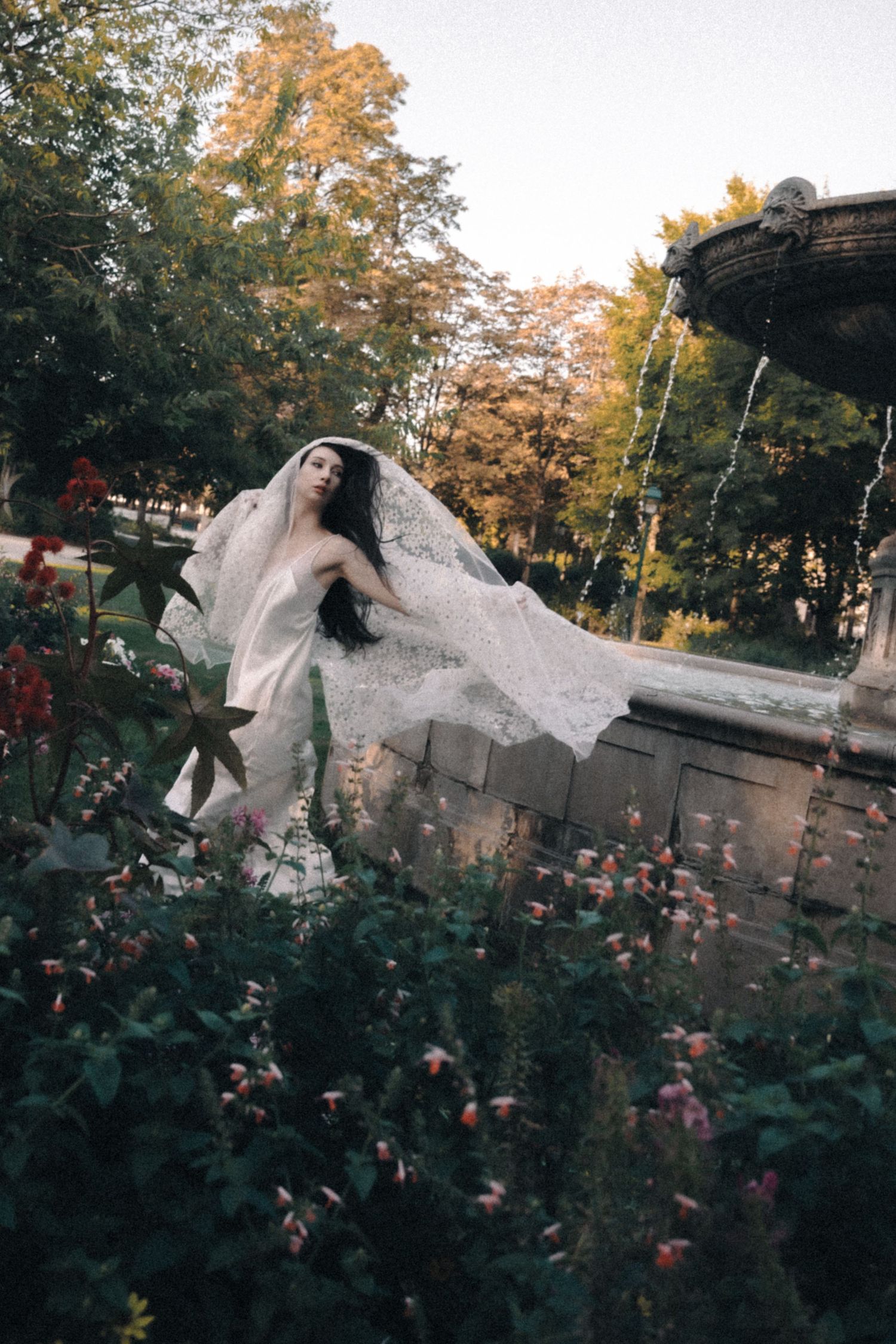 Bride in white wedding dress and flowing veil poses by stone fountain surrounded by blooming flowers in a lush garden.