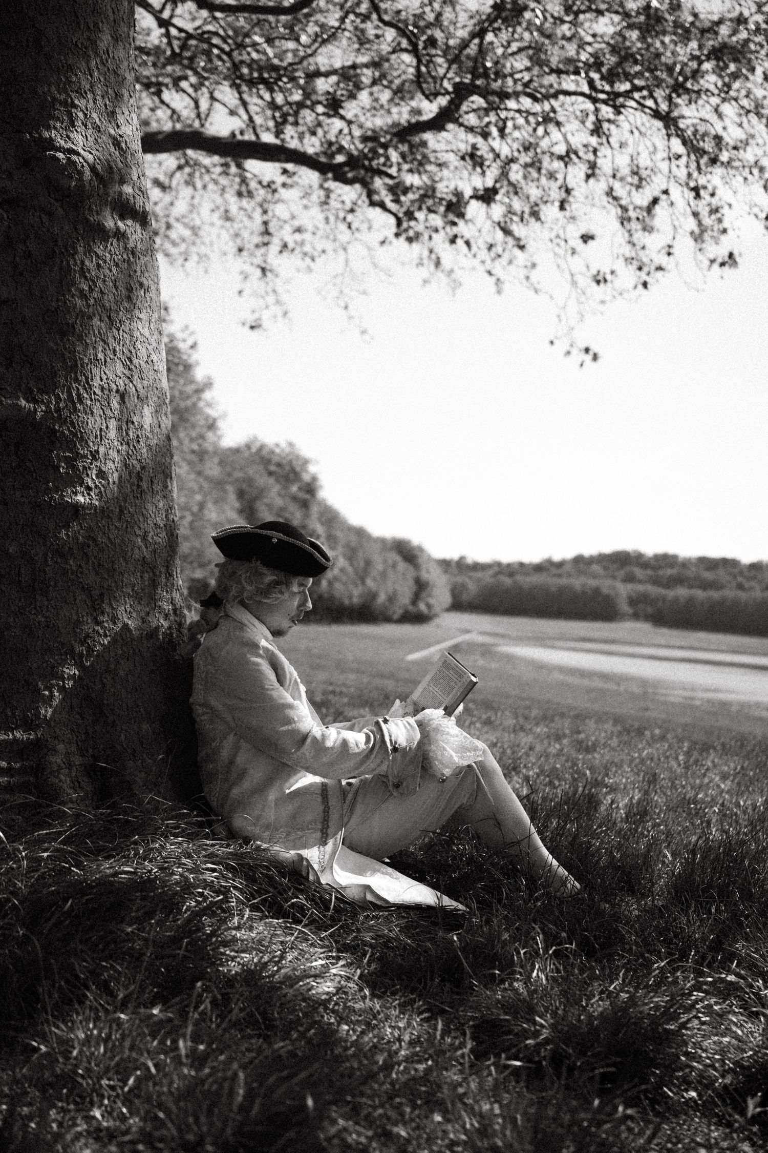 Black and white photo of a woman reading under a large tree in a peaceful countryside landscape.