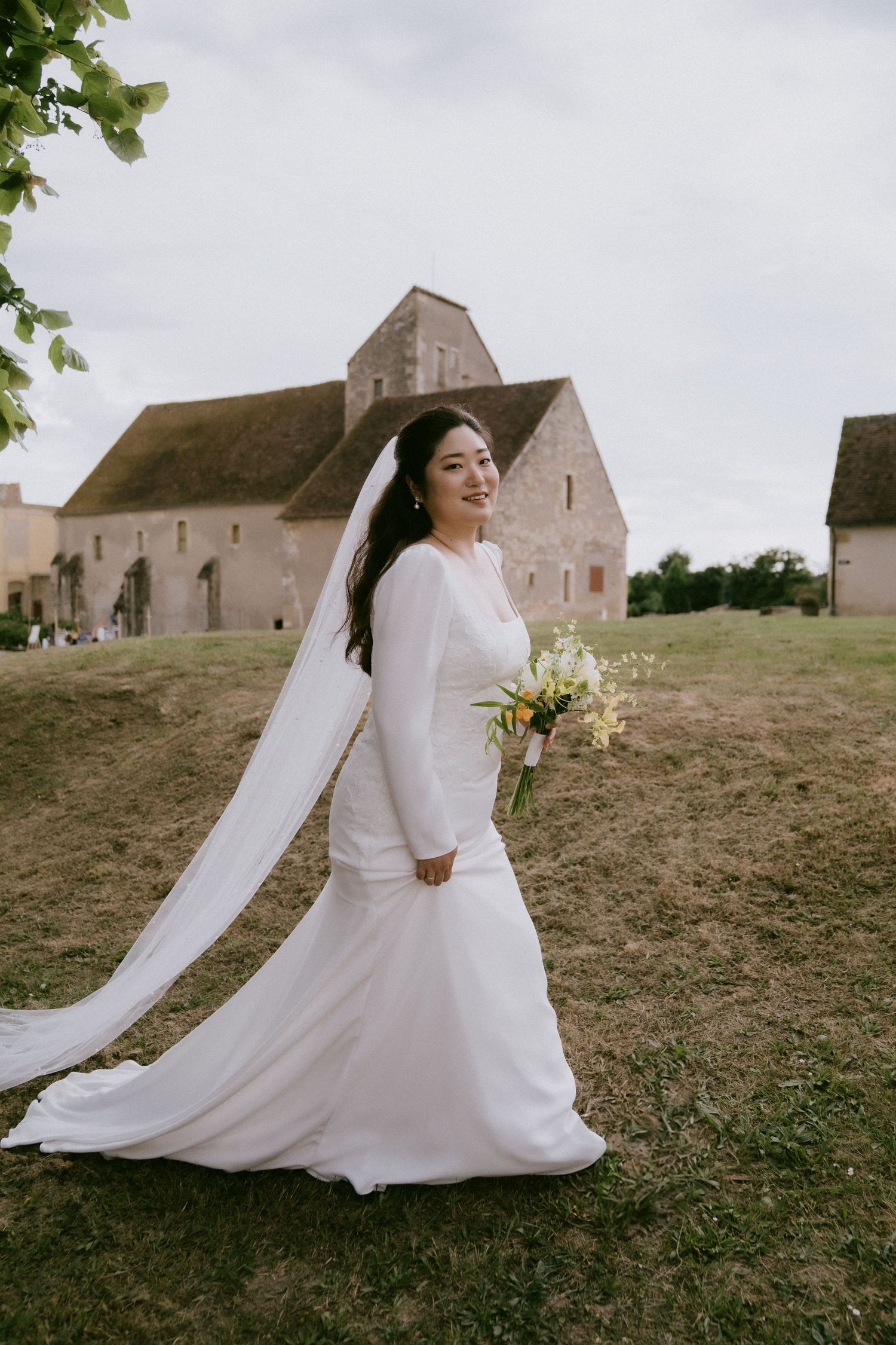 Bride in elegant white gown with flowing veil walks near historic stone church building on overcast day.