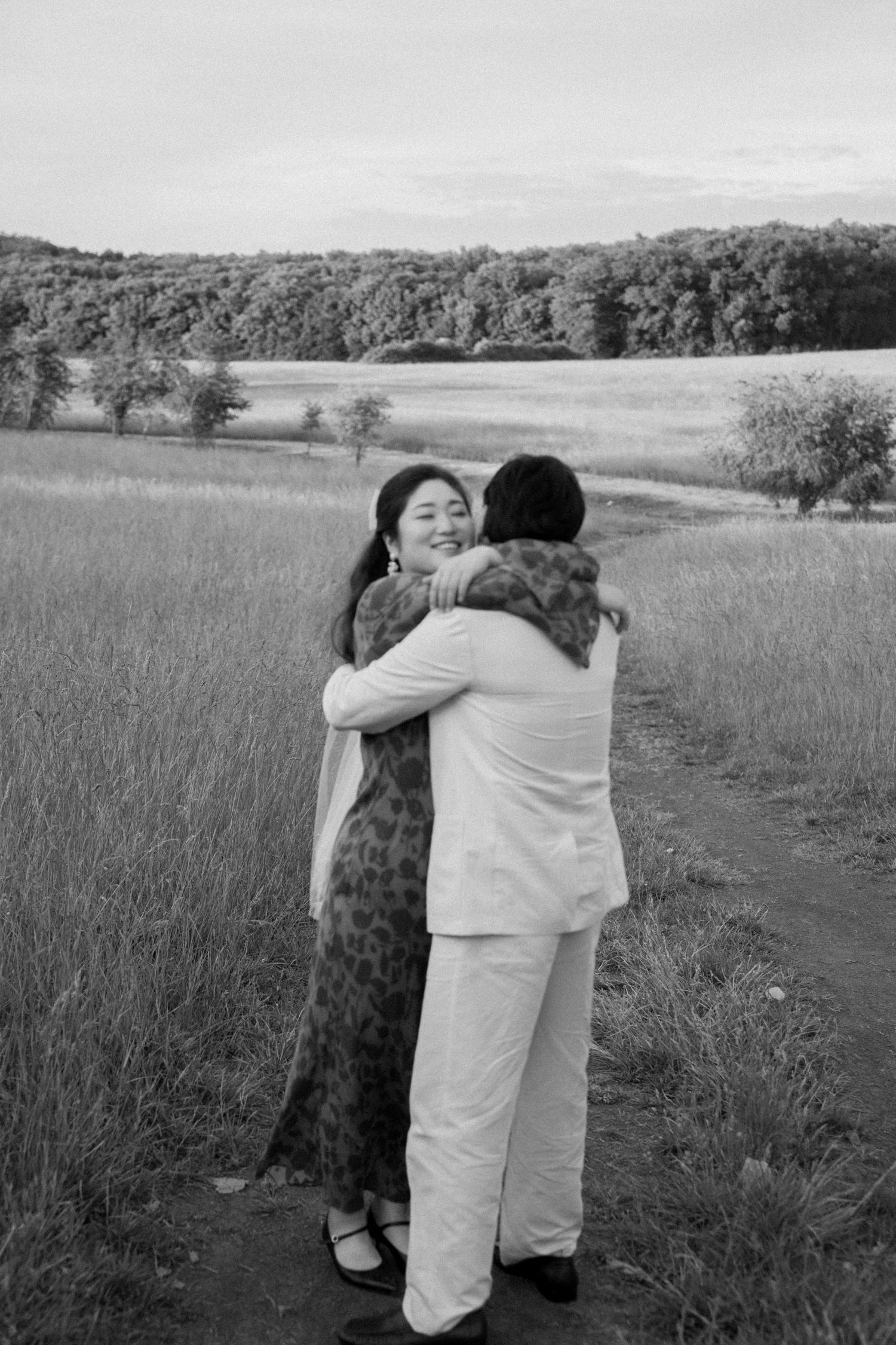 Two people embrace on a path in a scenic meadow landscape.