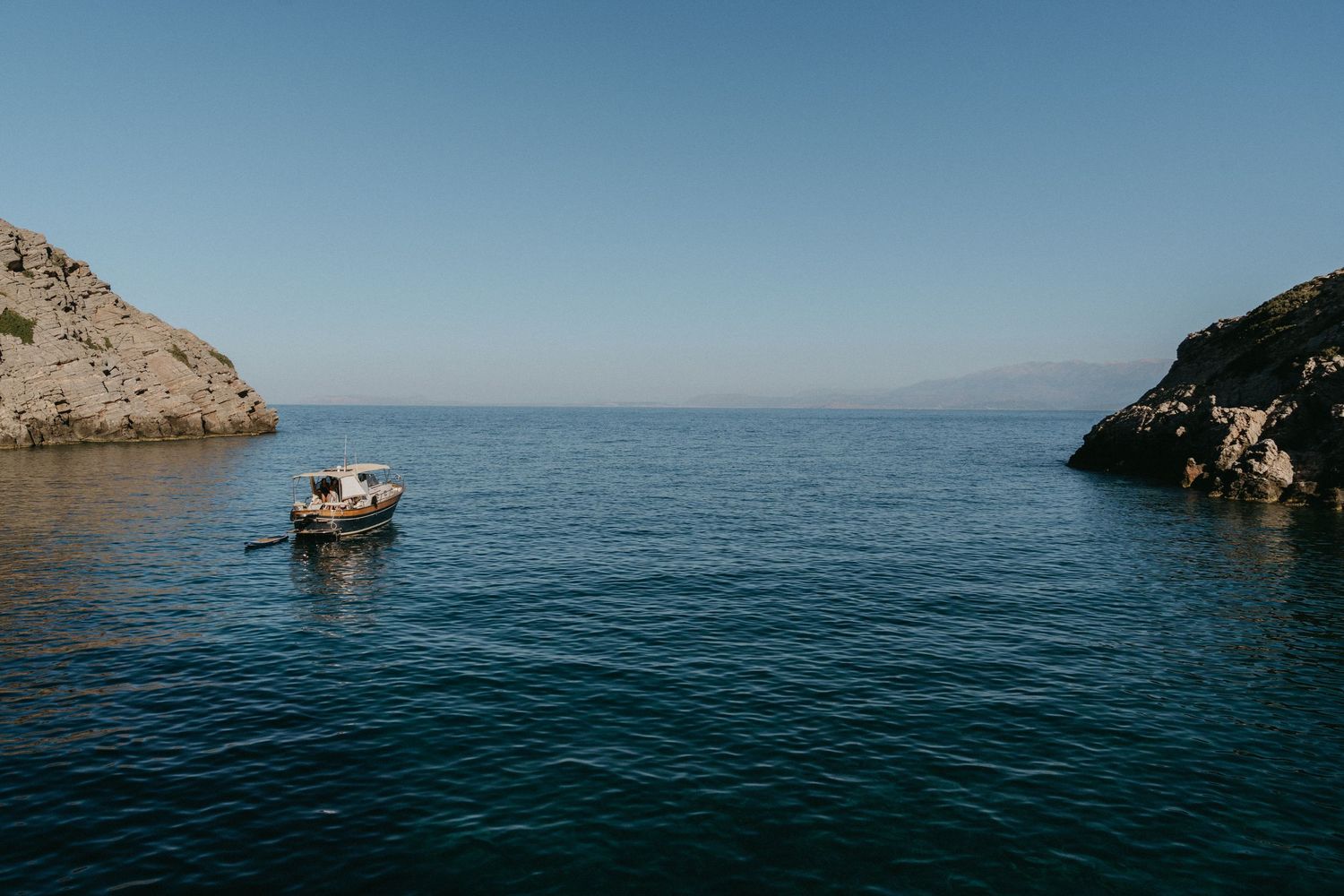 Scenic view of a small boat sailing near rocky coastal cliffs on a calm blue ocean in Crete, Greece.