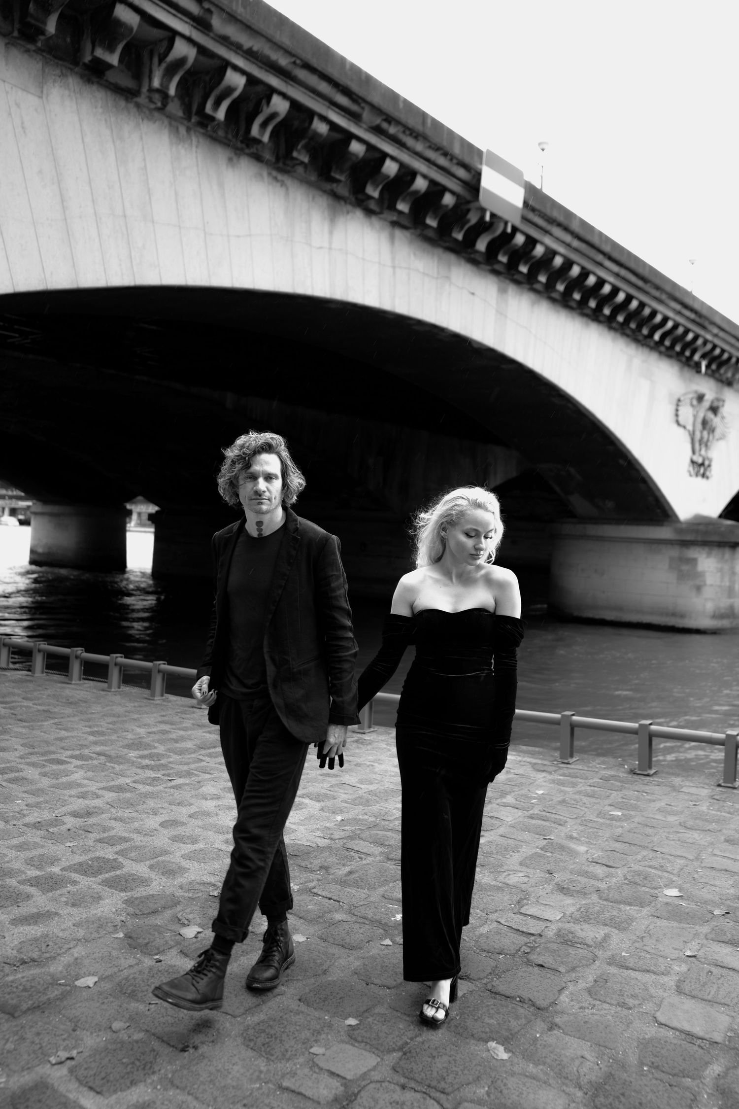 Editorial couple walking by the Seine in Paris, unposed and cinematic in timeless black outfits and soft natural light.