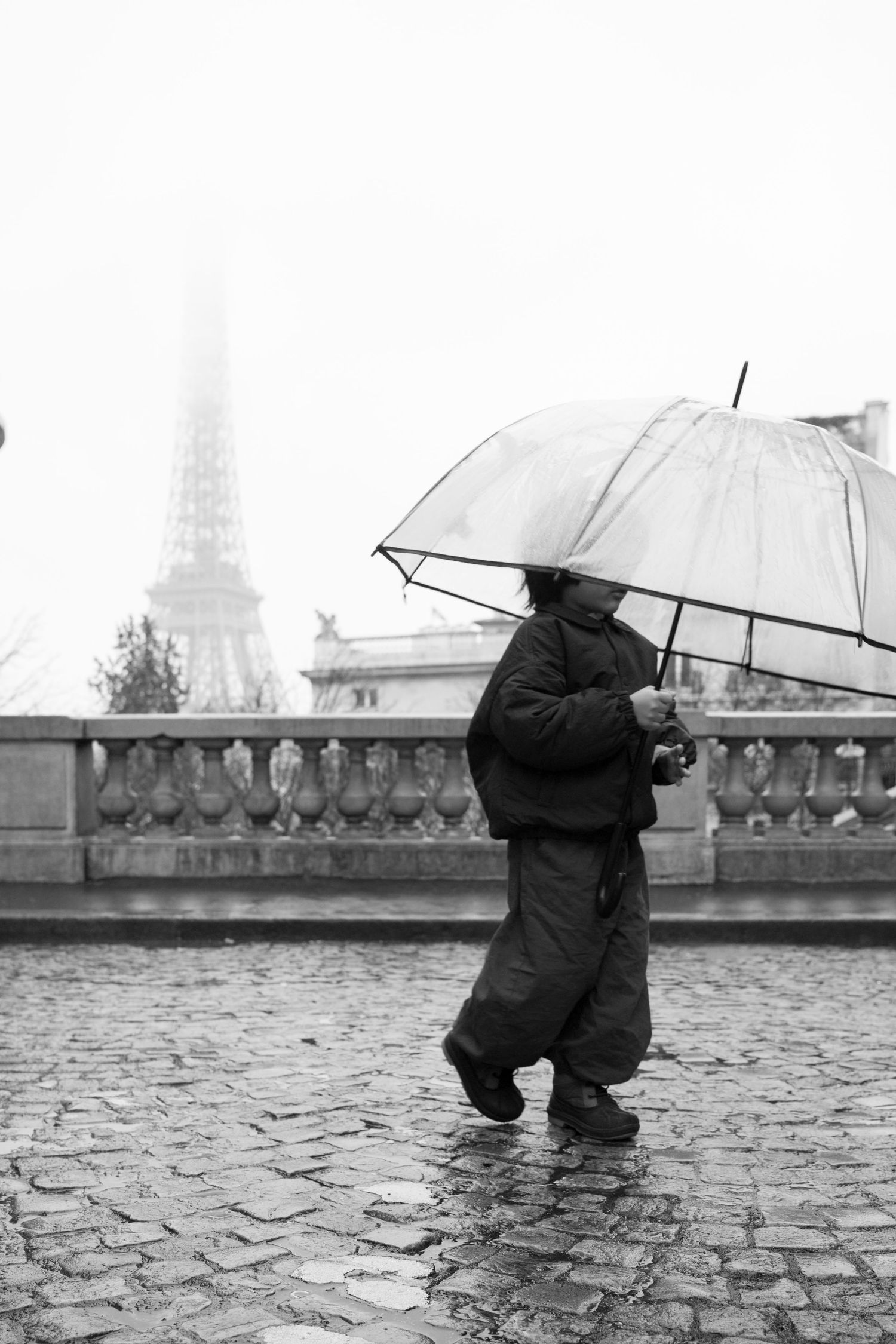 Person walking with clear umbrella on wet cobblestones in Paris, Eiffel Tower visible through fog, black and white photo.