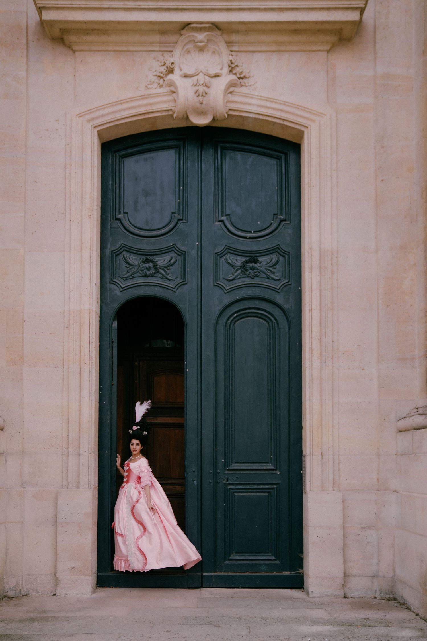 A person in a pink vintage ballgown stands in a grand doorway with ornate dark green doors and stone archway.