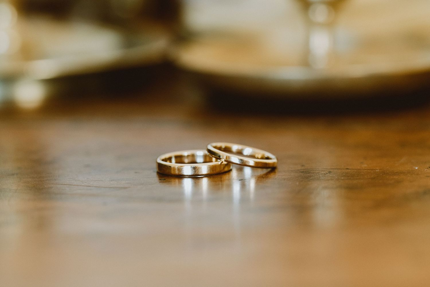 Gold wedding rings sit on a wooden surface with soft bokeh lighting in the background.