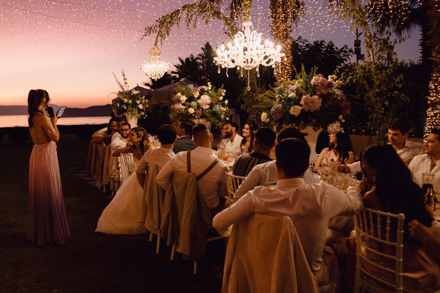 Wedding guests gather on a sunset terrace overlooking the water during an elegant outdoor reception celebration.