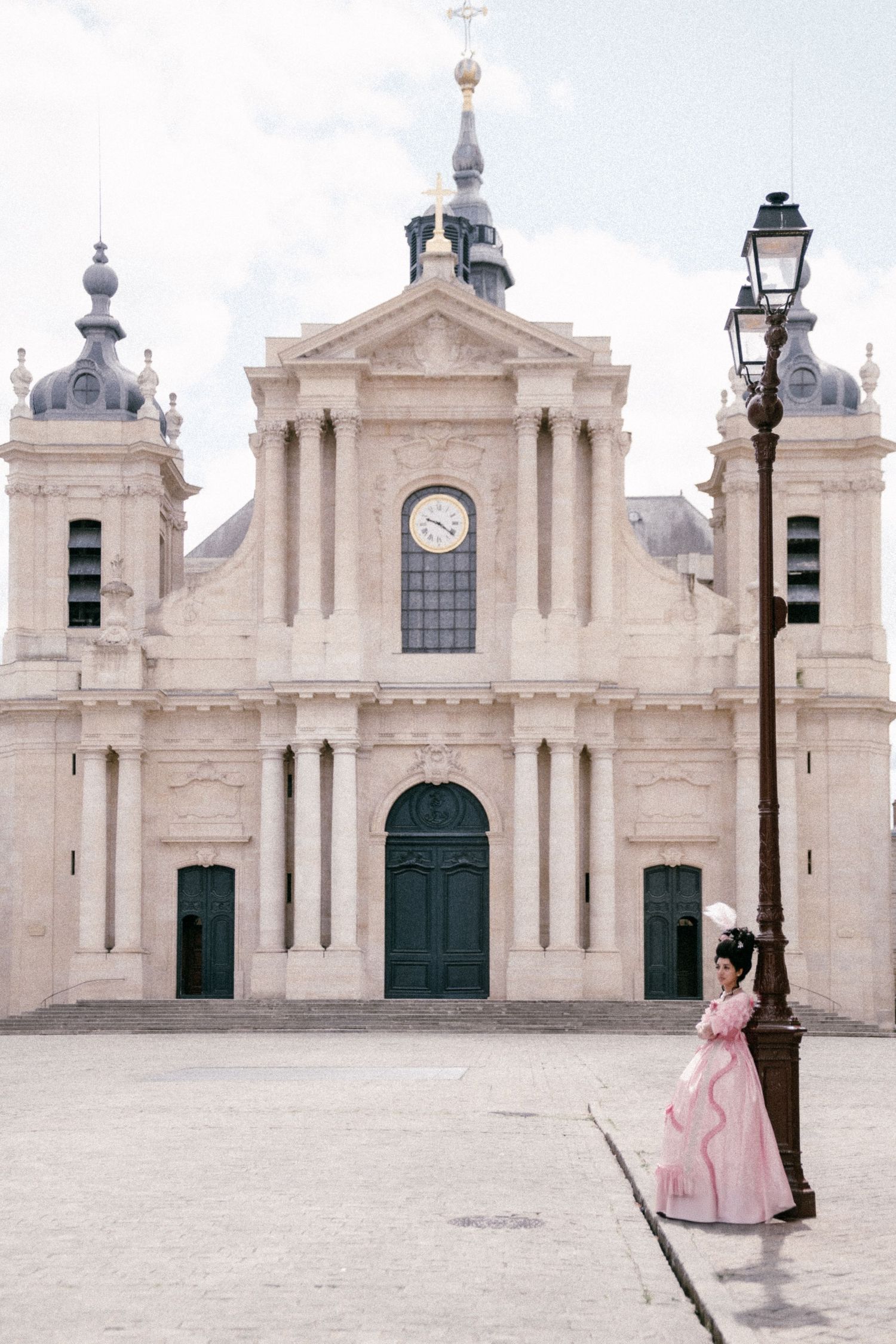 A person in a pink dress stands in front of a white baroque church facade with green doors and a clock tower.