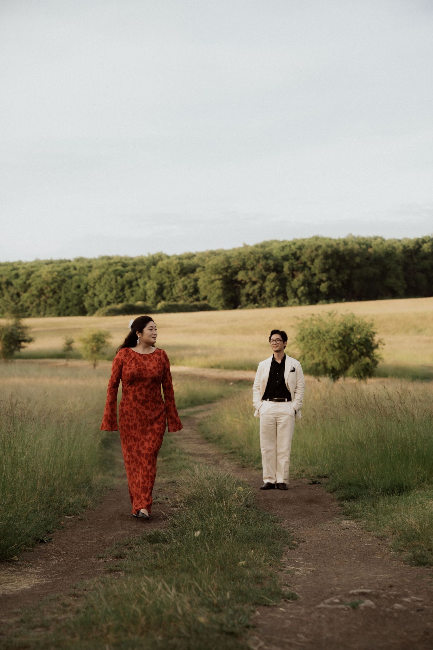 Couple walking through a grassy field at sunset, one wearing a flowing red dress.