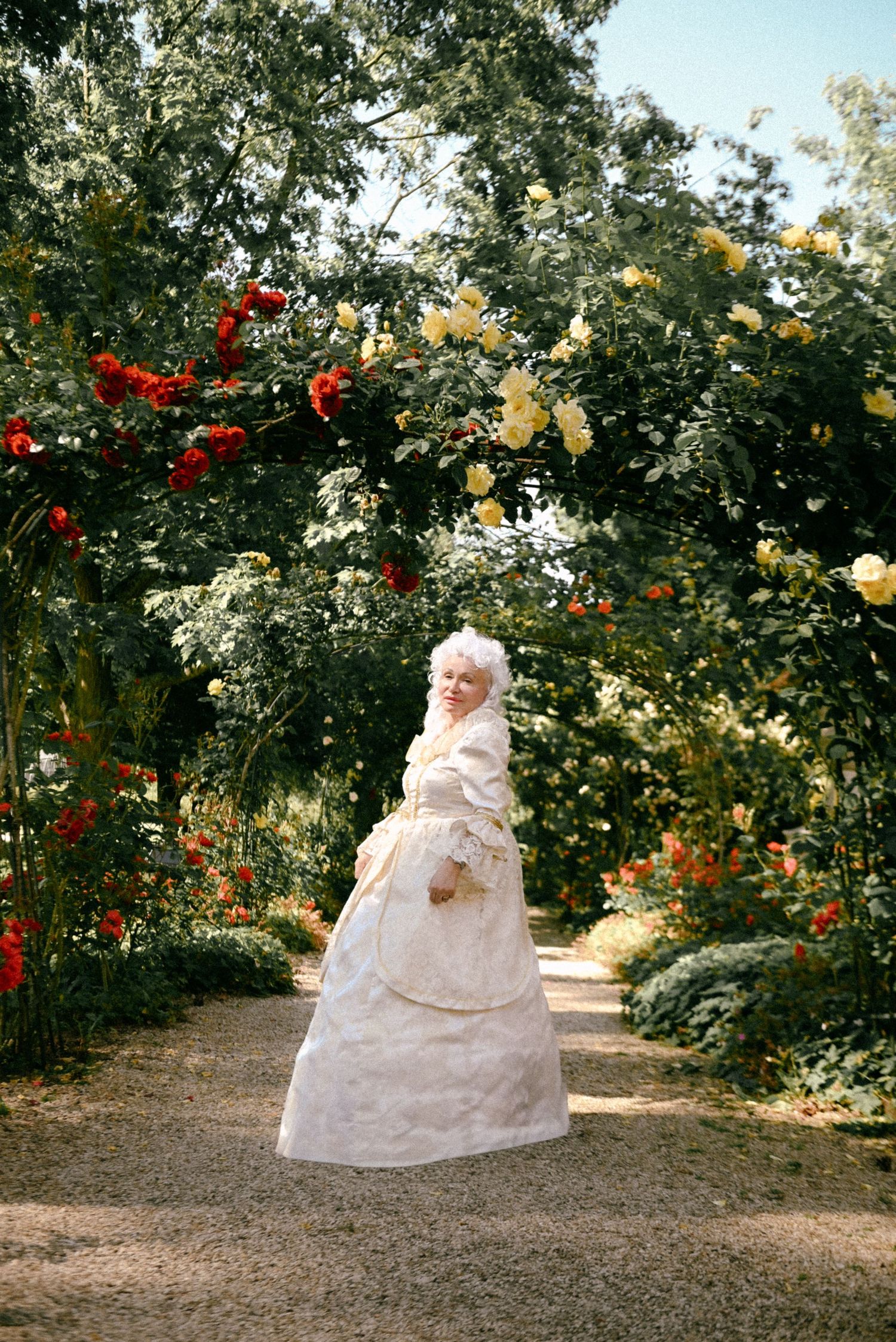 A person in a flowing white gown sits gracefully in a lush garden setting with blooming red and yellow flowers.