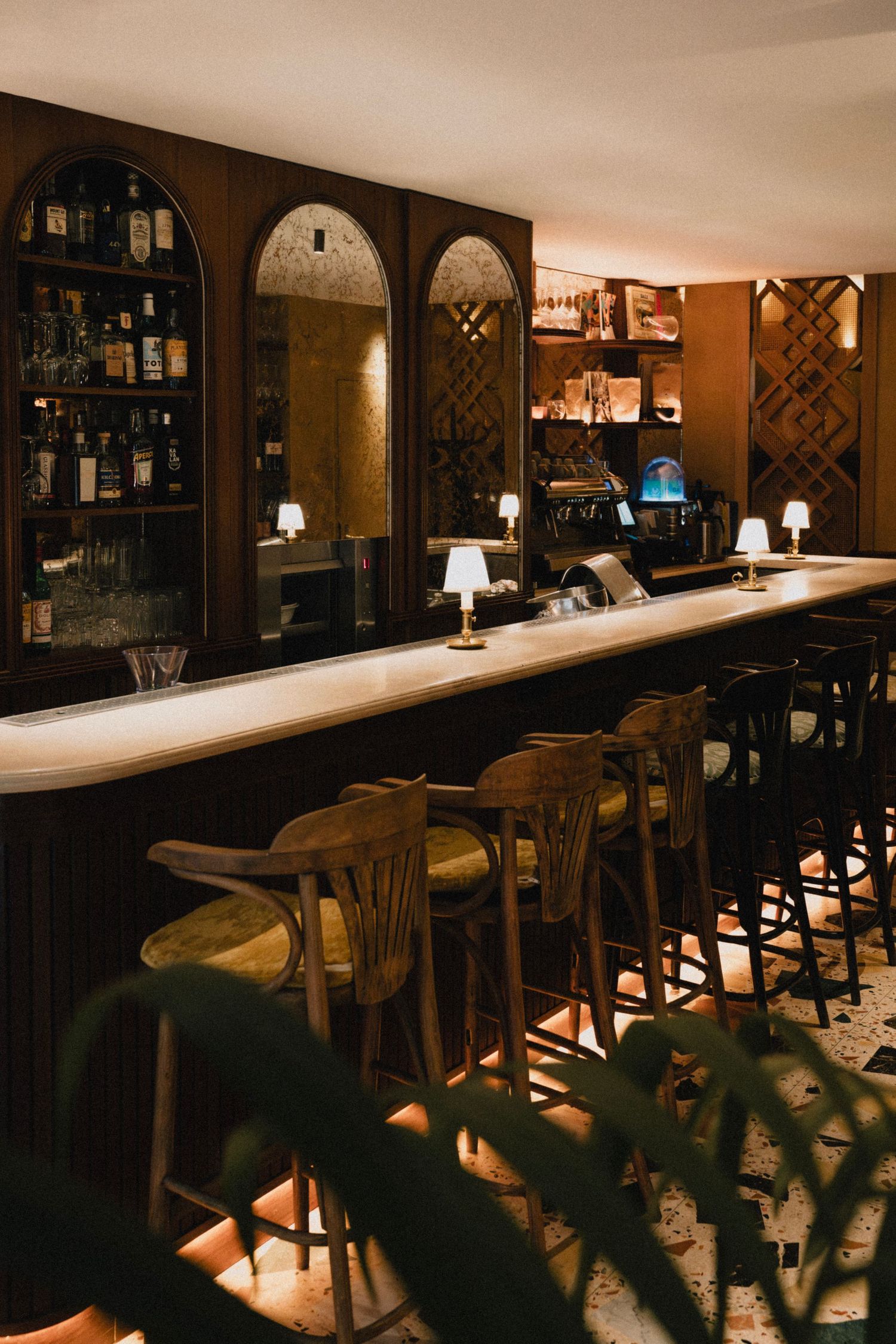 Dimly lit bar with wooden stools along a white countertop and bottles displayed on shelves in the background.