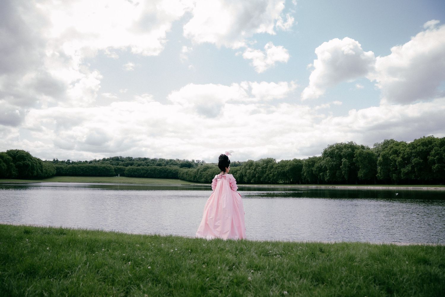 A person in a flowing pink gown stands at the edge of a serene lake surrounded by trees and clouds.