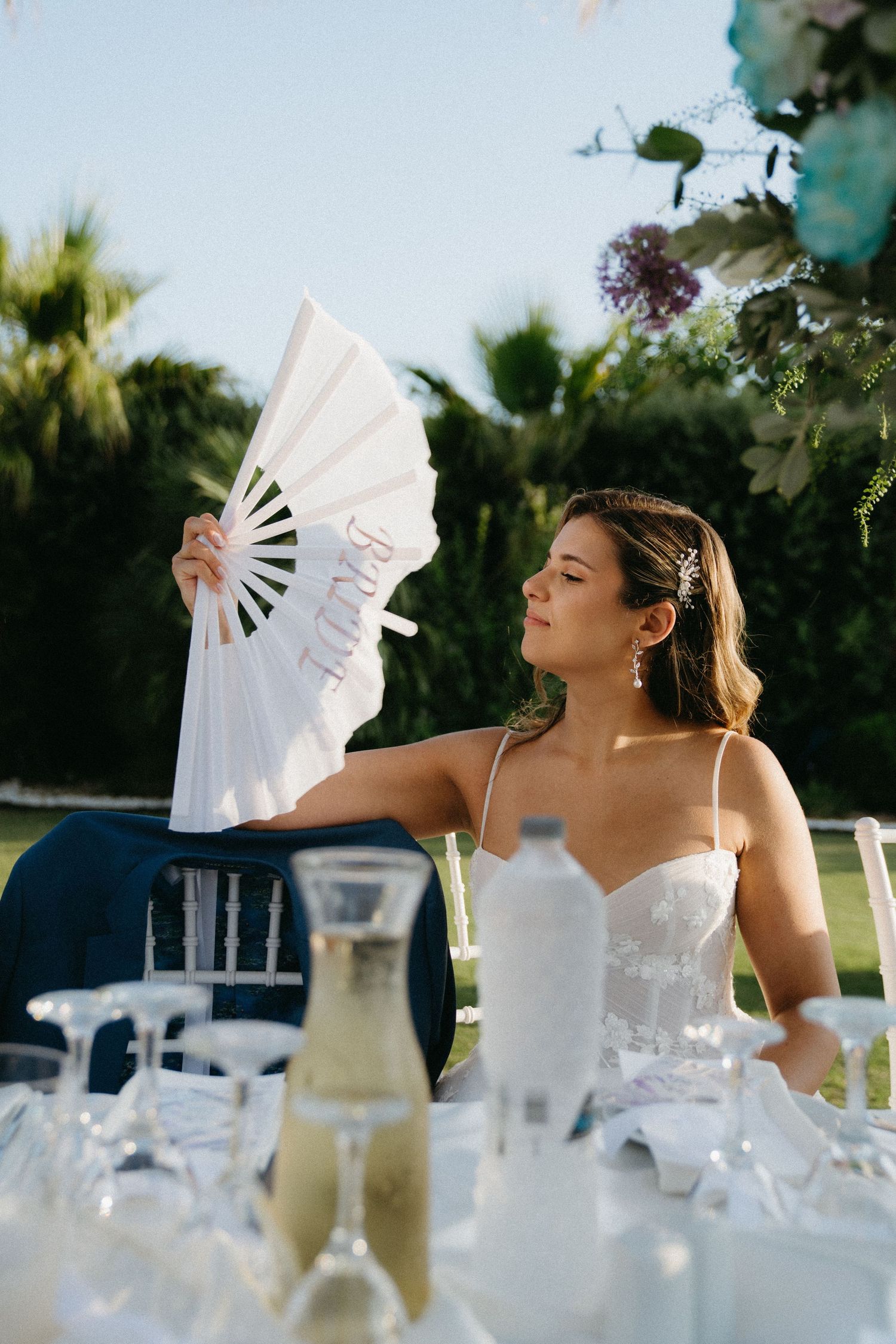 Wedding guest cools off with white paper fan at outdoor reception table during summer celebration.