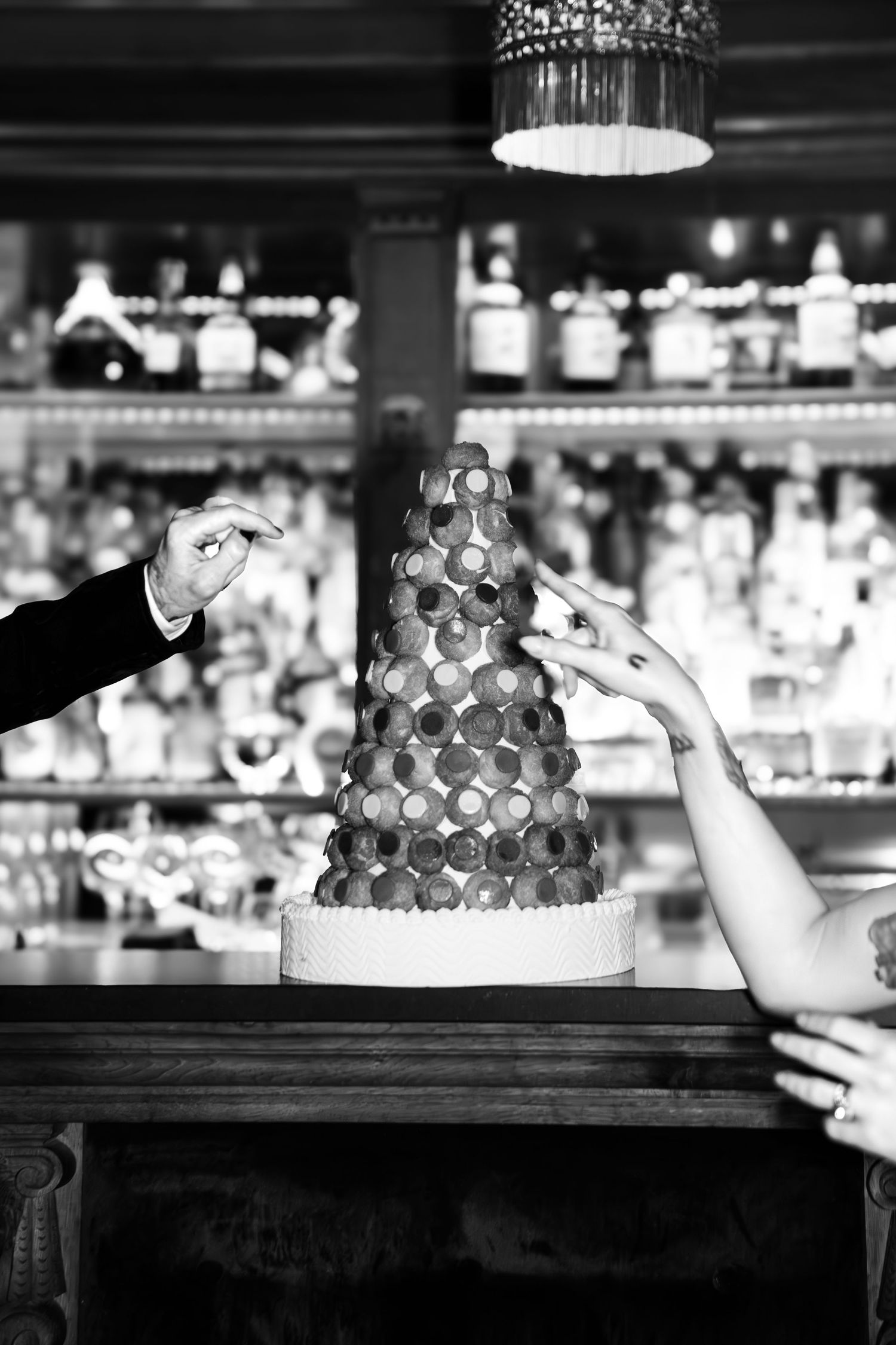 Black and white photo of hands decorating a tiered macaron tower dessert at a bar with bottles in background.