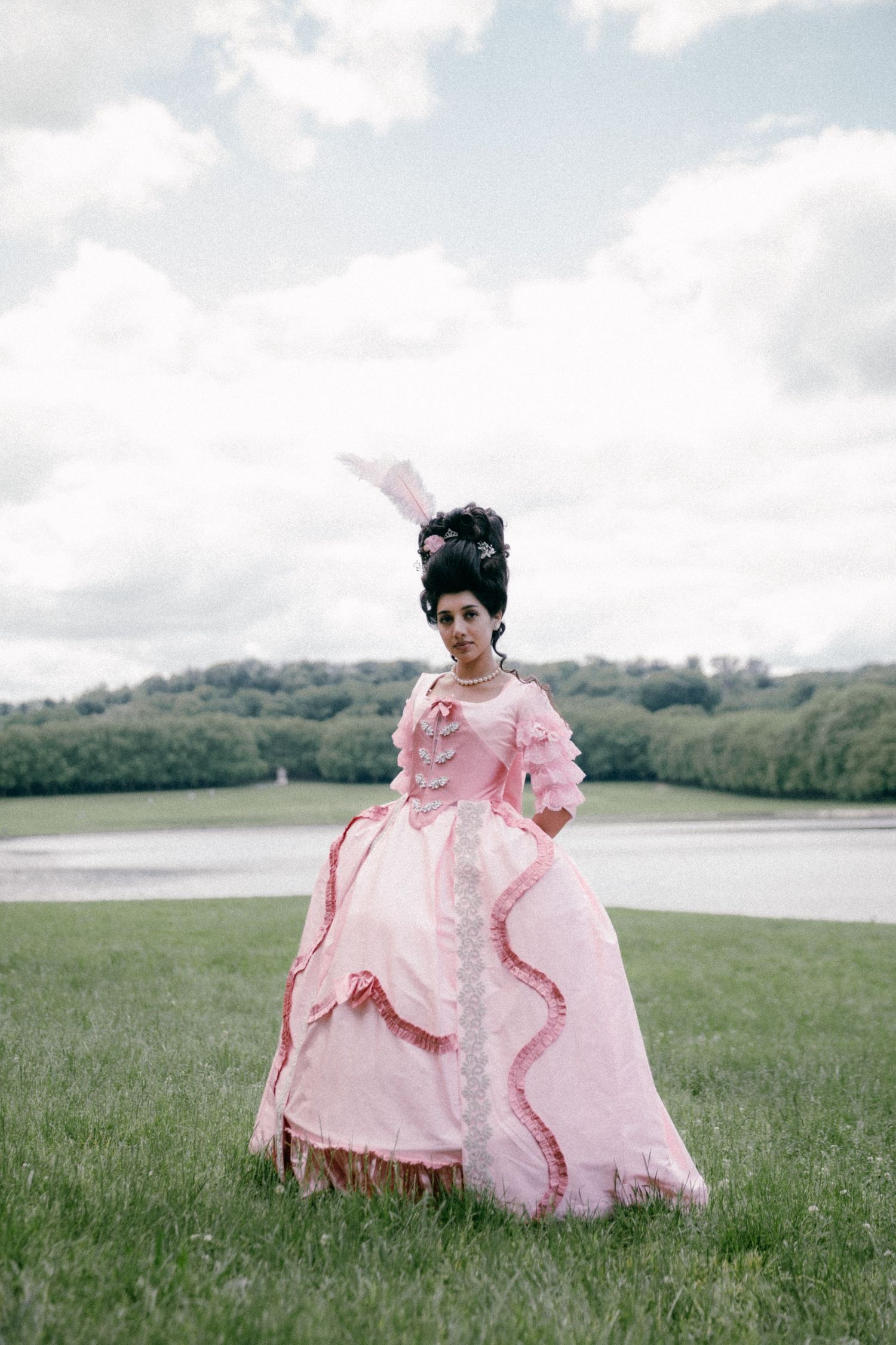 A person in a voluminous pink gown stands in a grassy field with a lake and trees in the background.