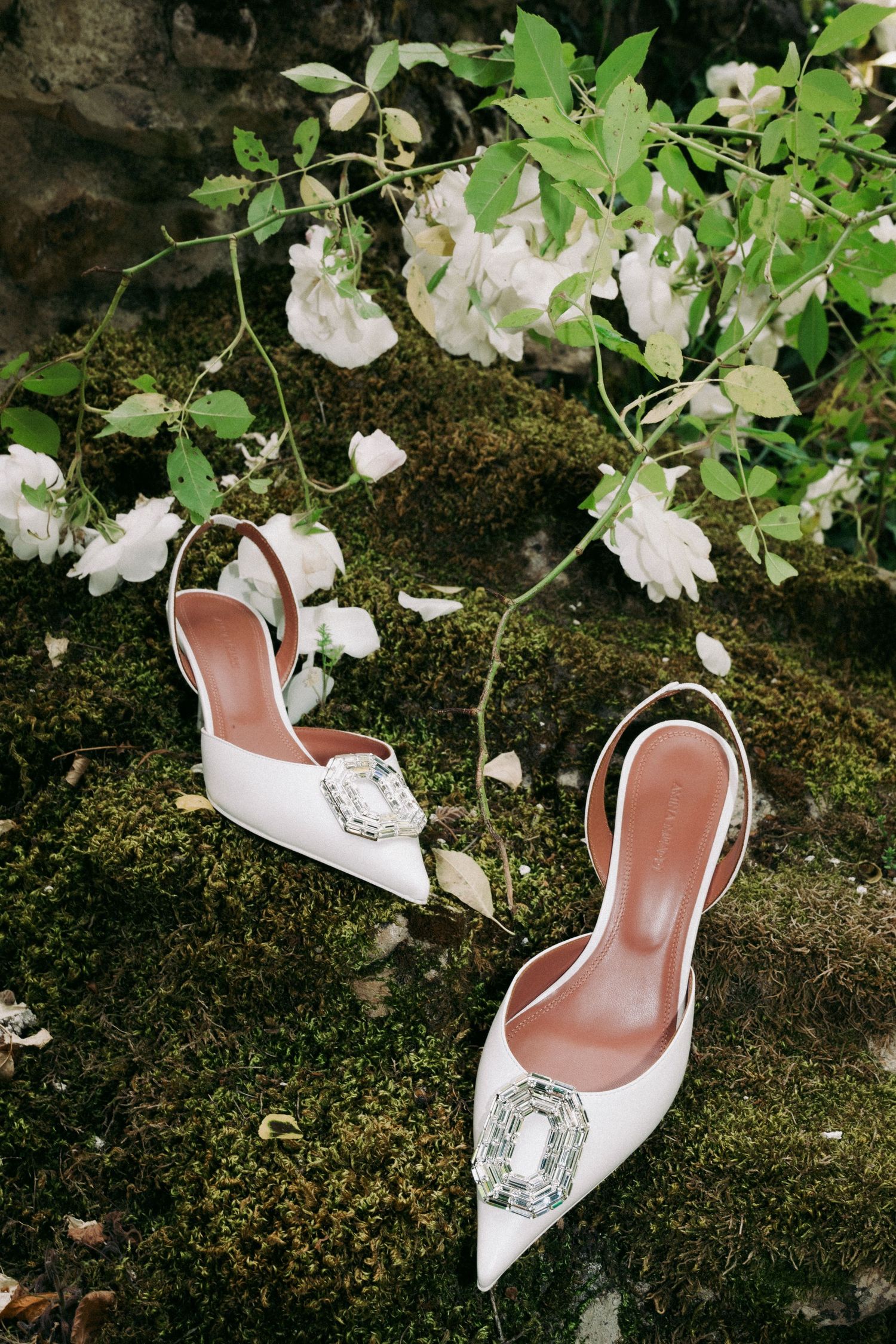 White bridal shoes arranged among delicate white flowers and green foliage on mossy ground.