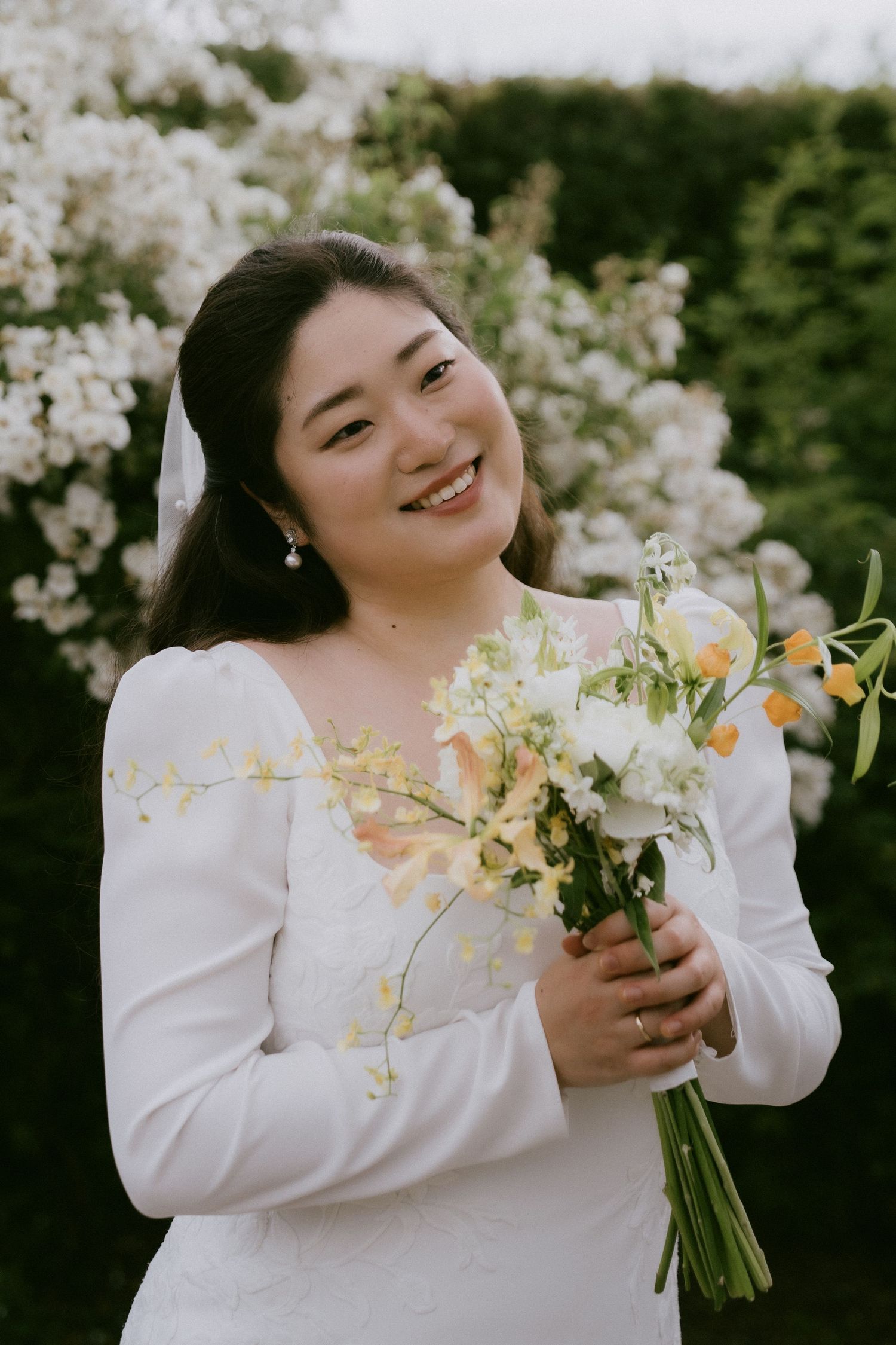 Bride in long-sleeved white wedding dress holds spring bouquet with white flowers against blooming garden backdrop.