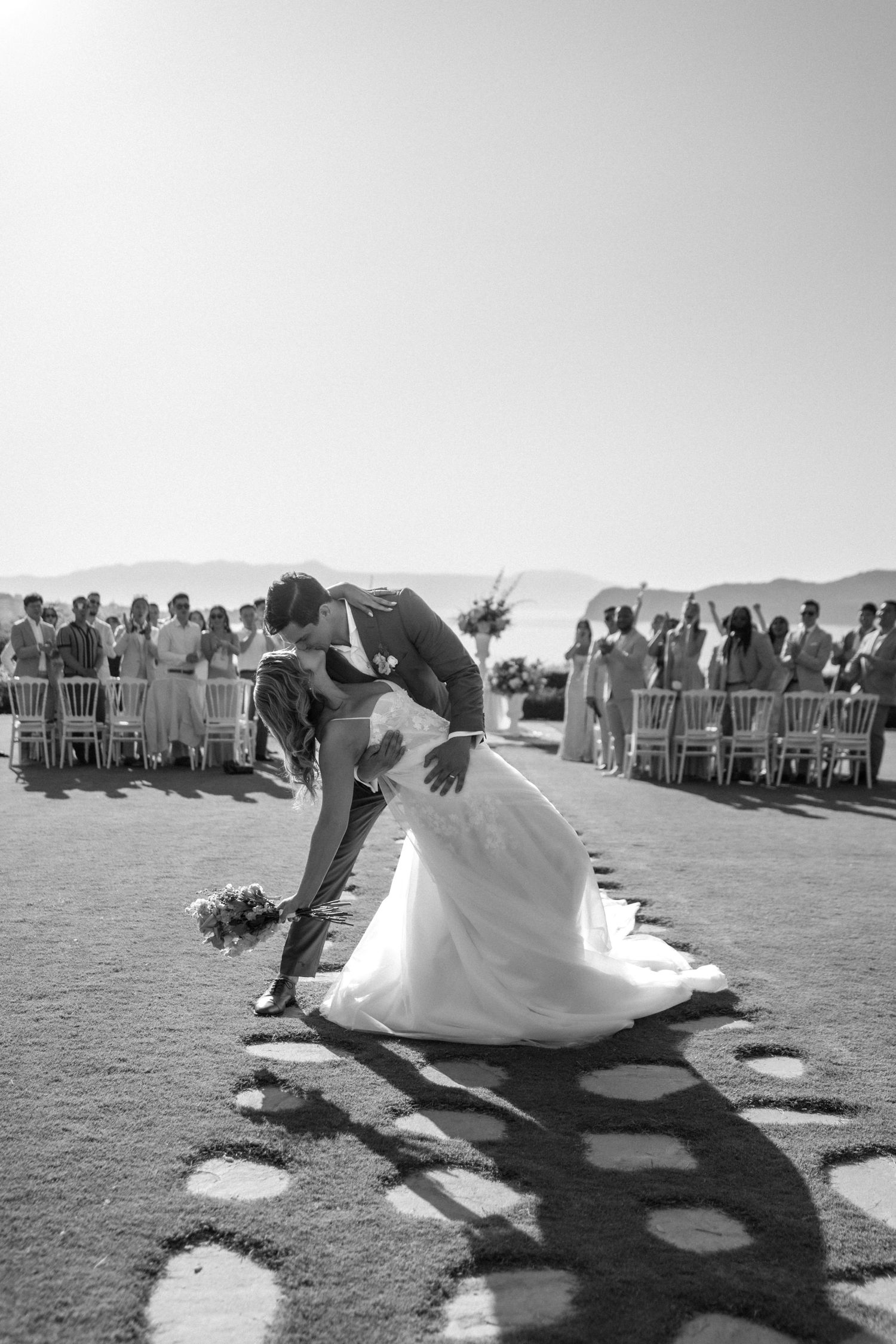 Wedding guests watch as a couple dips dramatically during an outdoor ceremony, creating a striking silhouette against the grass.