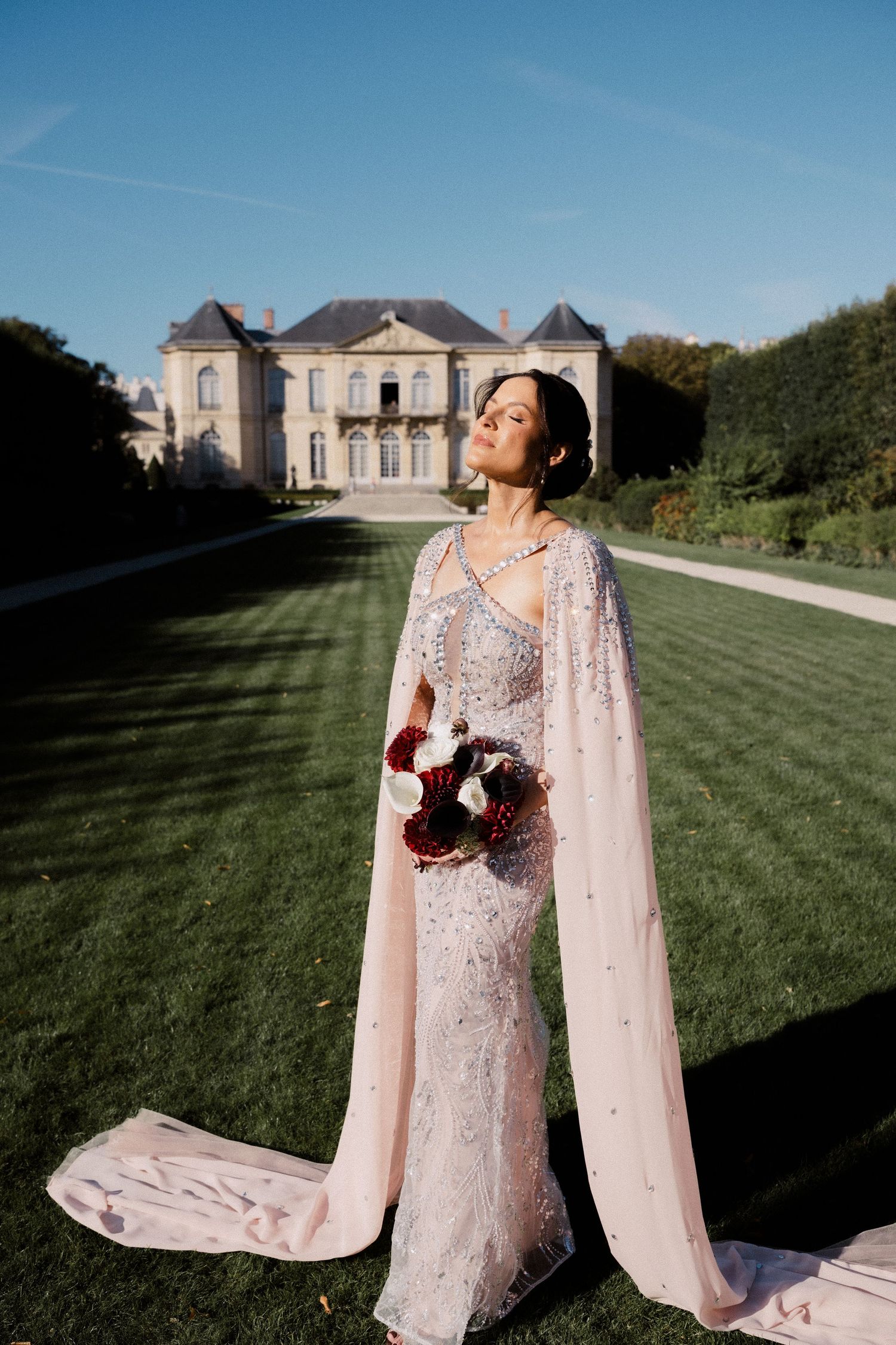 Bride in beaded blush gown with cape holds bouquet before grand French chateau on sunny day.