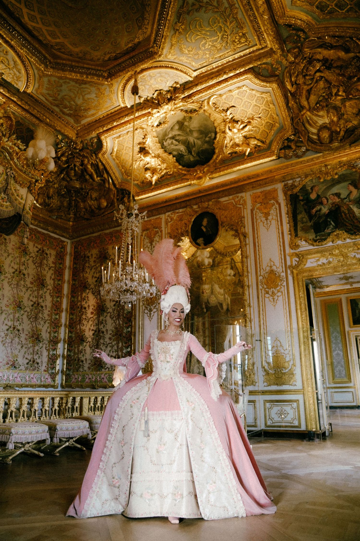 A figure in an ornate pink and white baroque gown stands in a luxurious palace room with gilded walls and chandeliers.