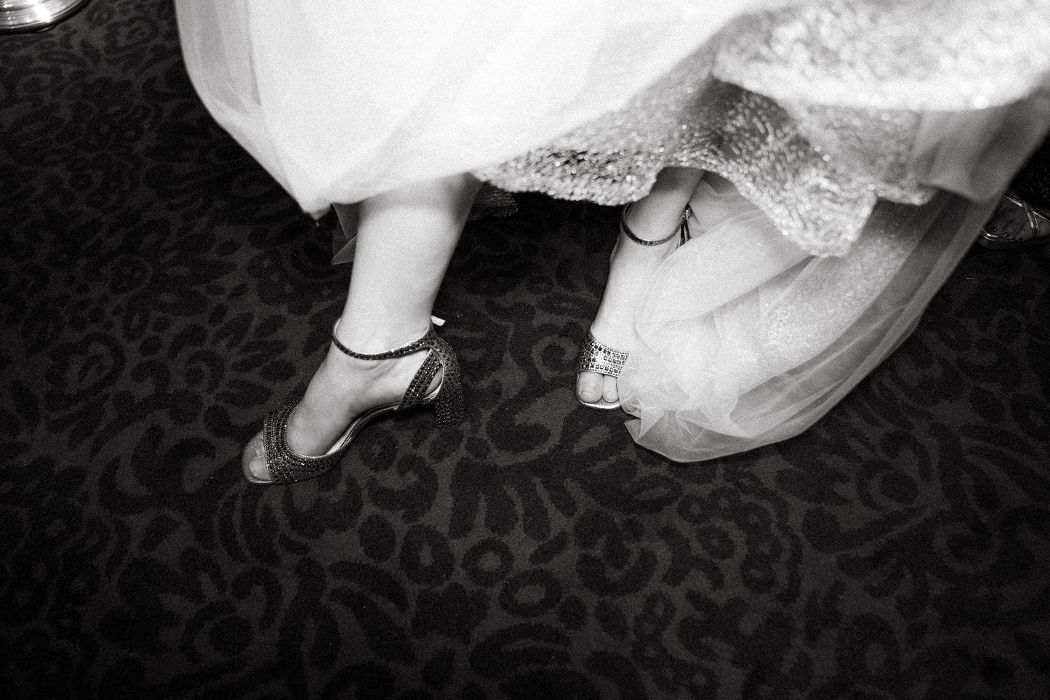 Black and white photo of elegant bridal shoes and dress hem against a patterned dark carpet.