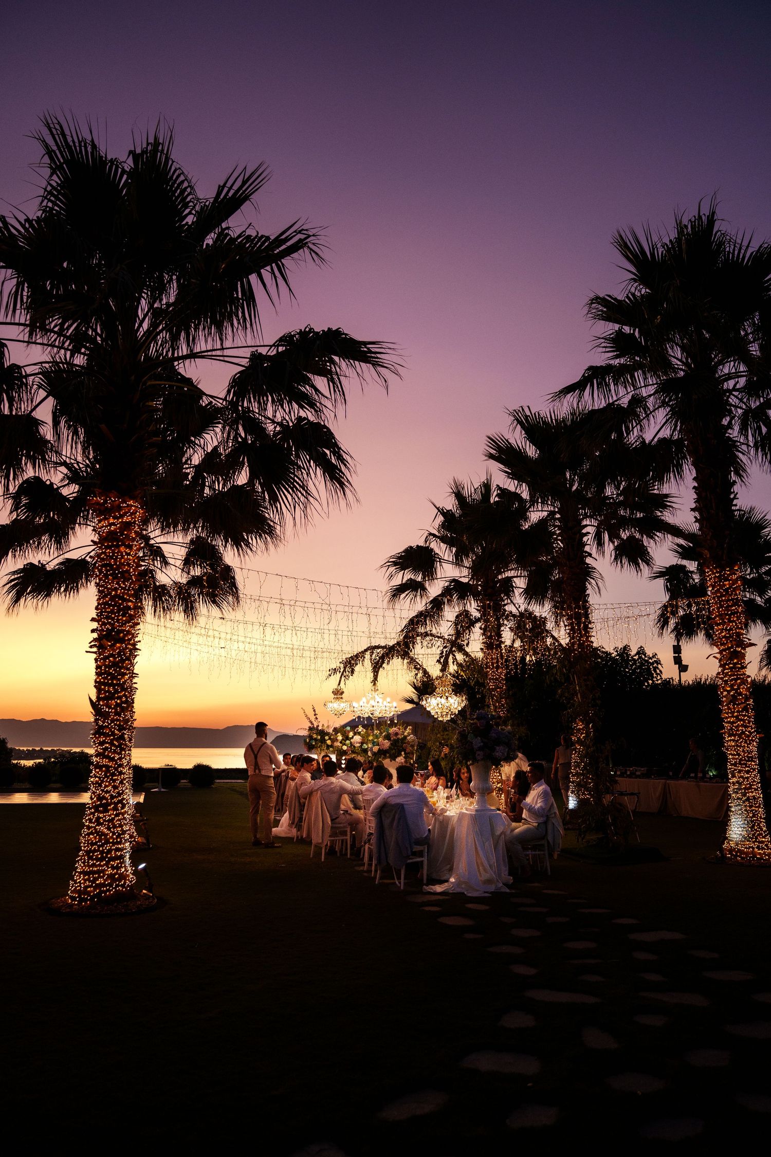 Palm trees silhouetted against a purple and orange sunset sky with string lights creating a romantic outdoor ambiance.
