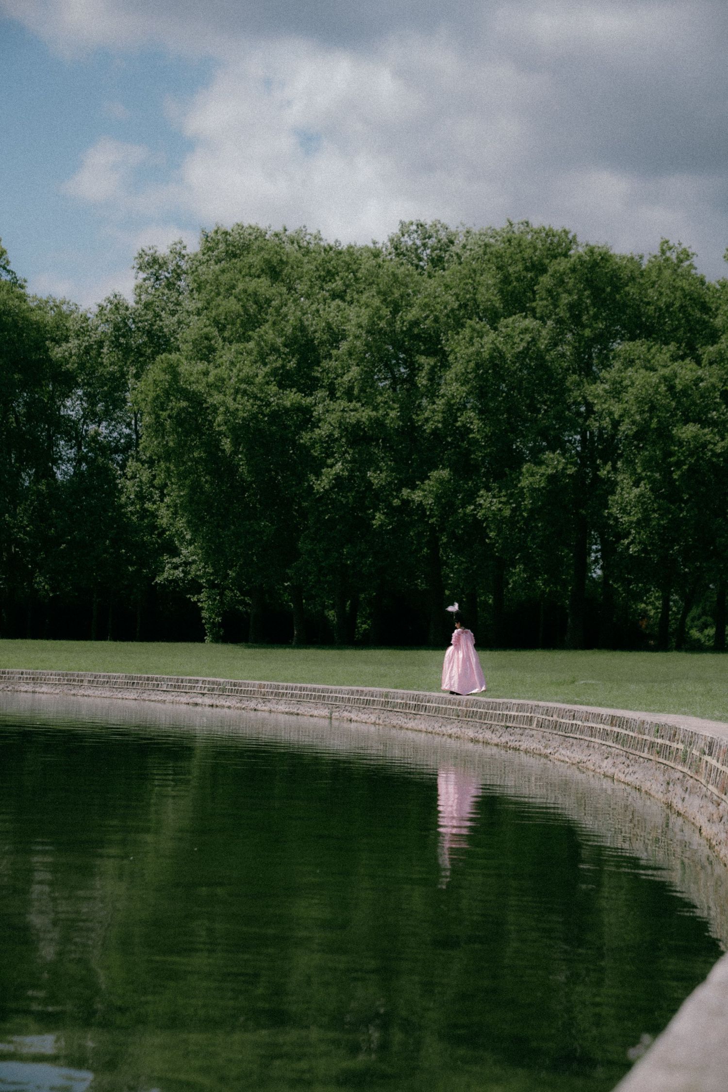 A lone figure in pink reflects in dark water along a curved stone wall under cloudy skies.