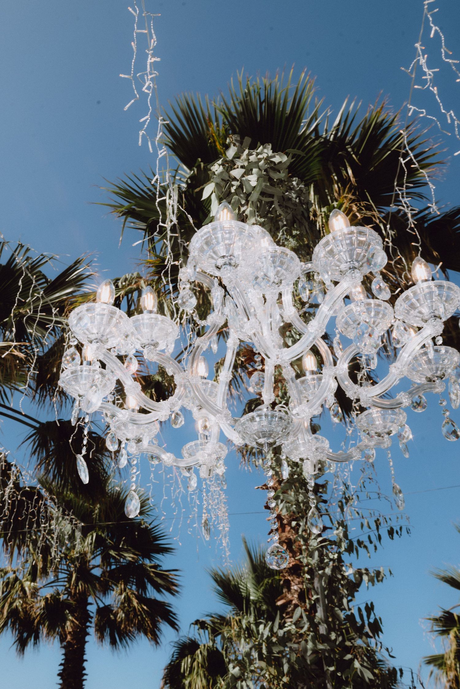 Delicate white paper decorations and palm trees create an elegant outdoor wedding ceremony setting against a blue sky.