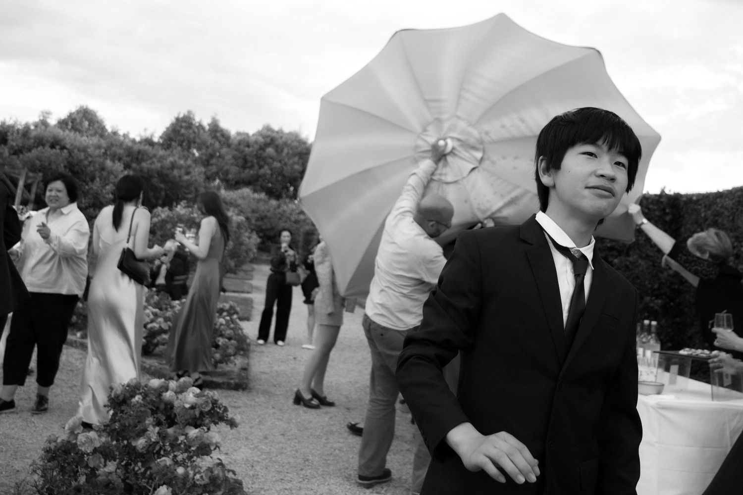 A person in a suit holds a white umbrella at an outdoor gathering in black and white photography.
